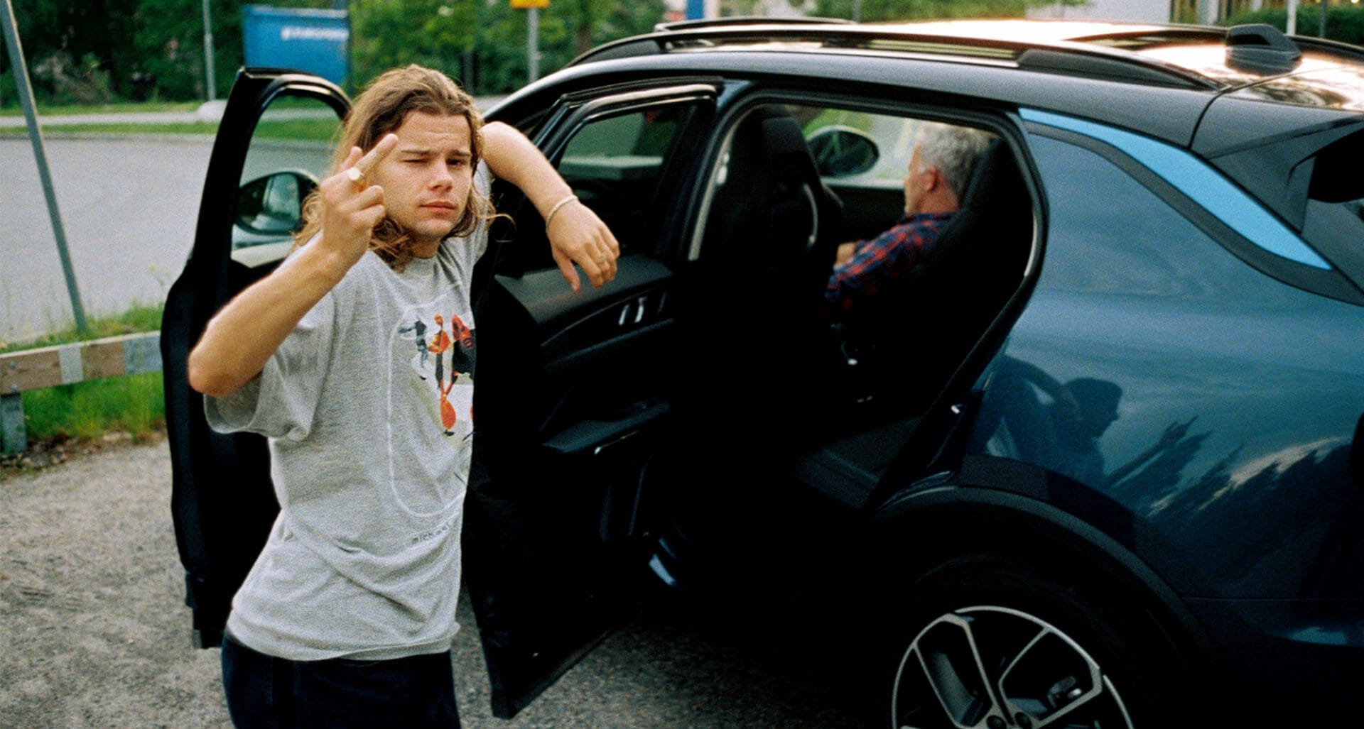 A young man with long wavy hair making a gesture with his middle finger, standing beside a dark blue car with the driver, an older man, inside wearing glasses and a checkered shirt, in an outdoor parking lot surrounded by greenery.