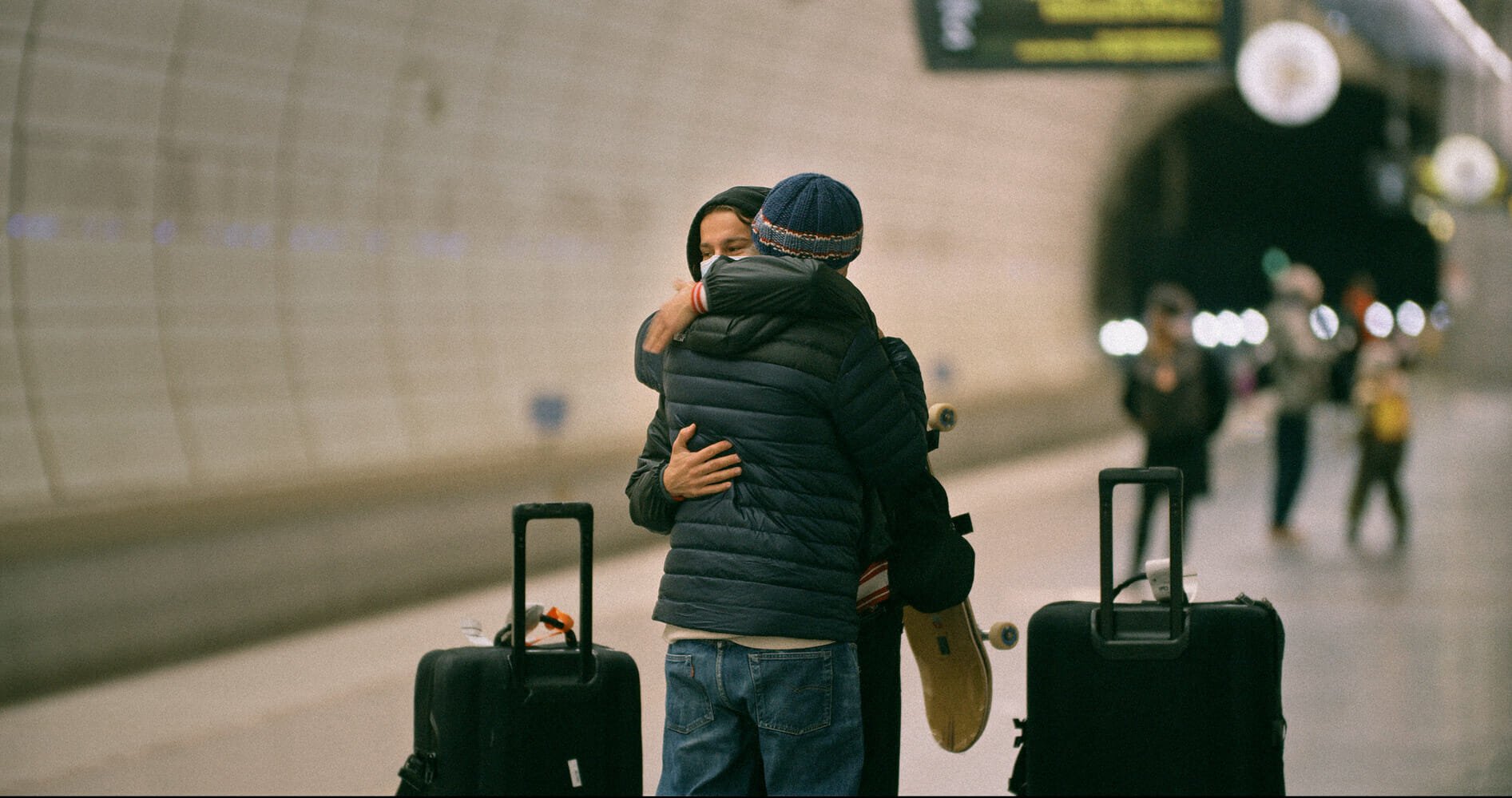 Two people hugging at an airport with luggage nearby, one holding a skateboard, others walking in the background.
