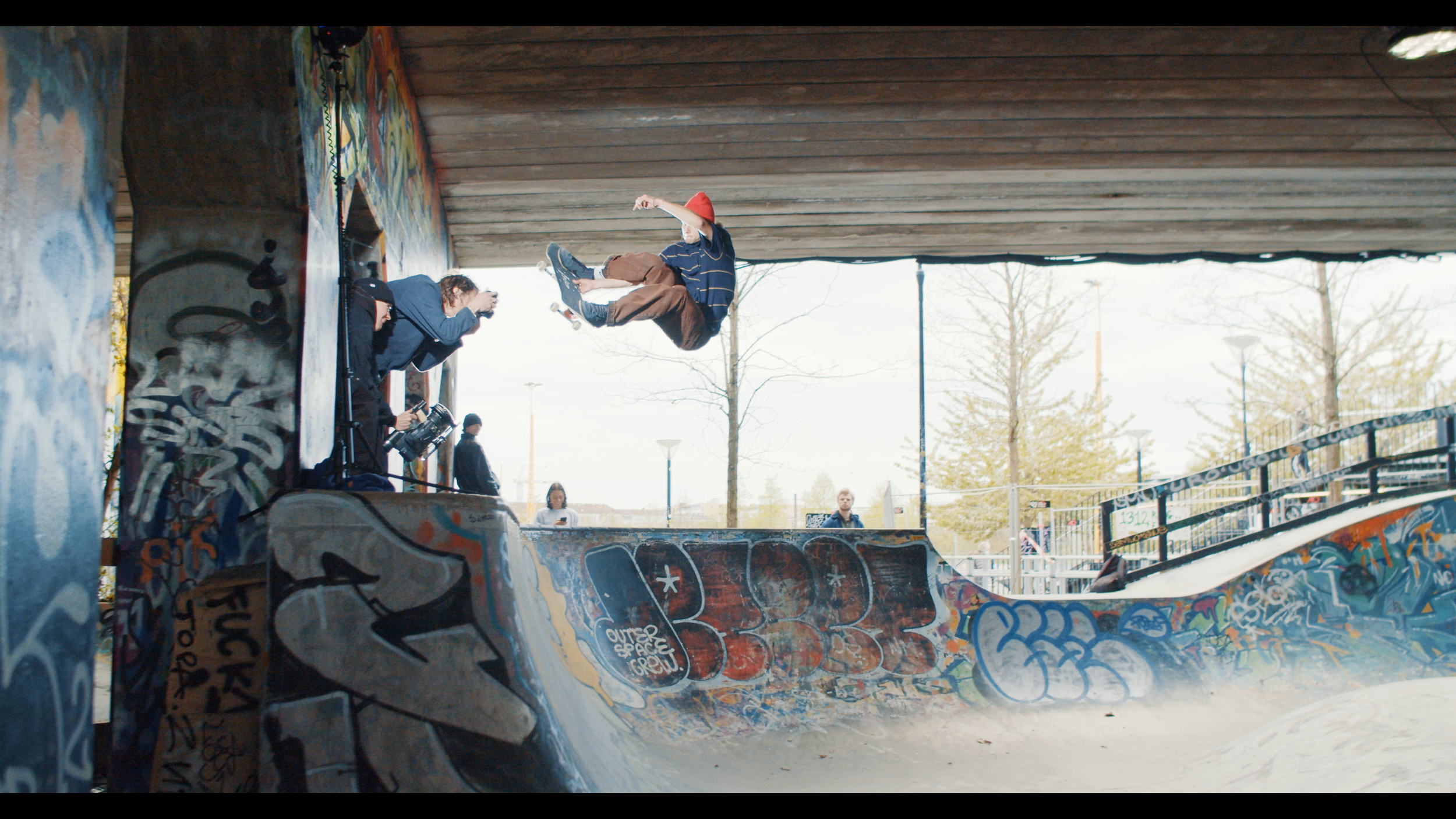 Skateboarder performing a trick in a graffiti-covered skate park with a wooden roof, while a photographer captures the moment and several people watch.