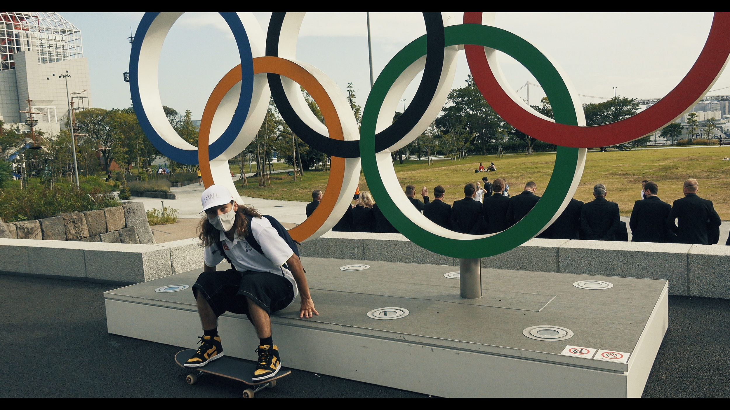 A young person wearing a white cap, face mask, and skateboard crouched by the Olympic rings sculpture at an outdoor park area.