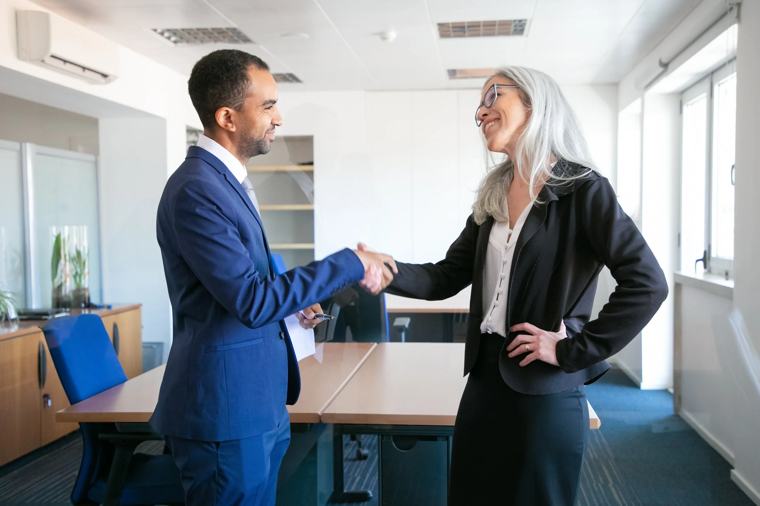 Homme en costume bleu serrant la main d'une femme en tailleur noir dans un bureau moderne