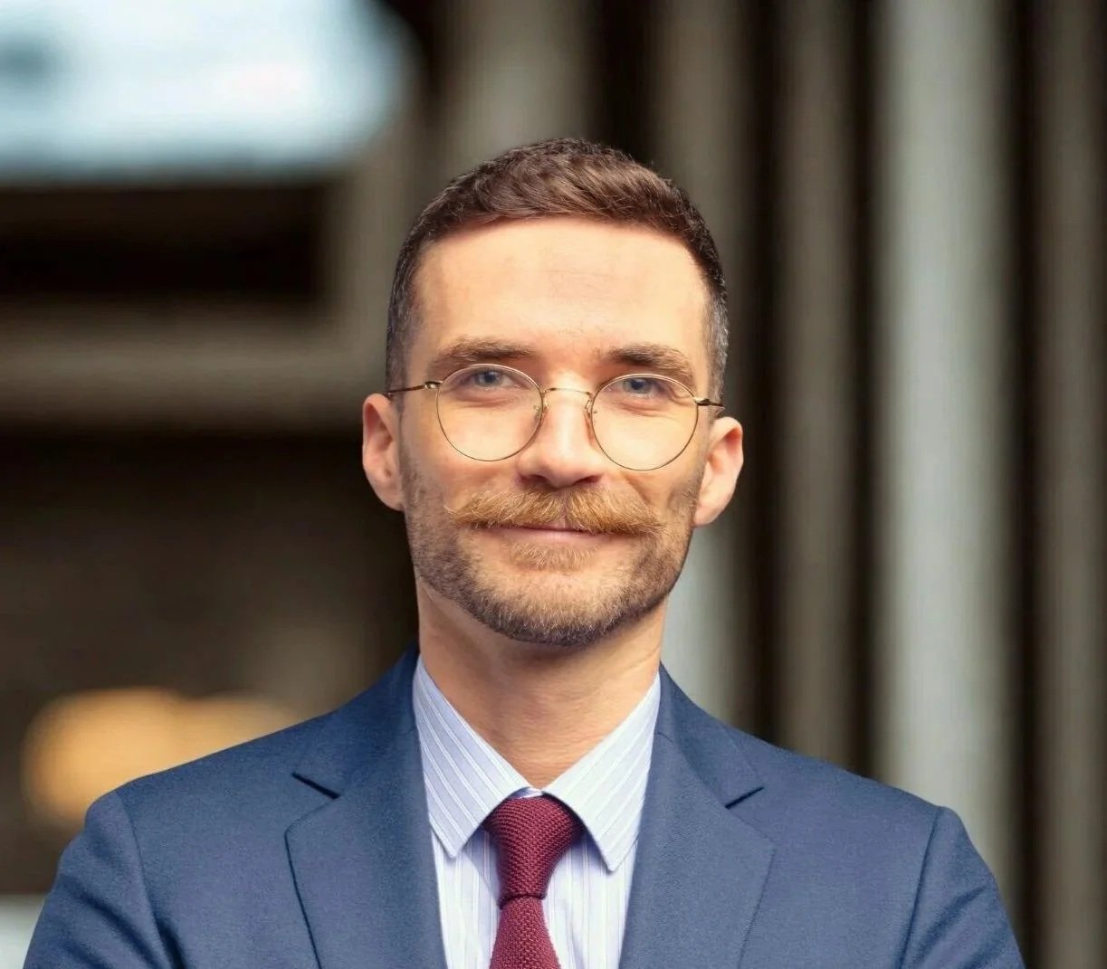 Portrait d'un homme d'affaires vêtu d'un costume bleu, portant des lunettes rondes, avec une barbe et une moustache, souriant devant un fond flou.
