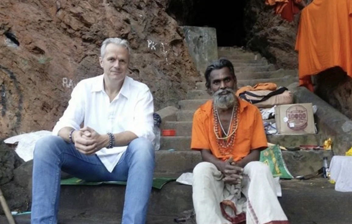 Two men sitting on stone steps against a rocky background, one Caucasian in a white shirt and jeans, the other South Asian in orange traditional attire with beads, with bags and clothing around them.