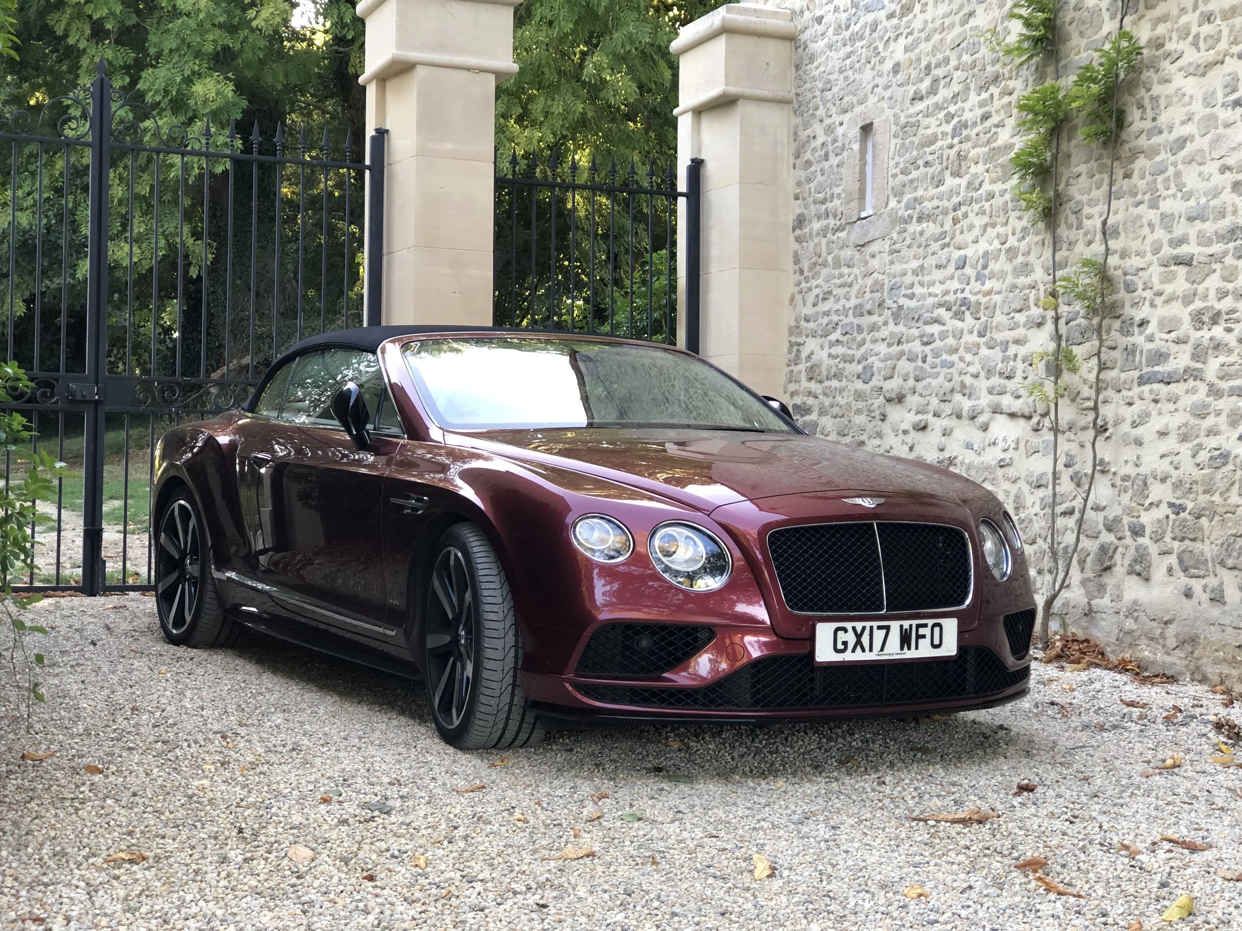 A maroon luxury convertible car parked on a gravel driveway in front of a stone wall and black wrought iron gate, with greenery in the background.