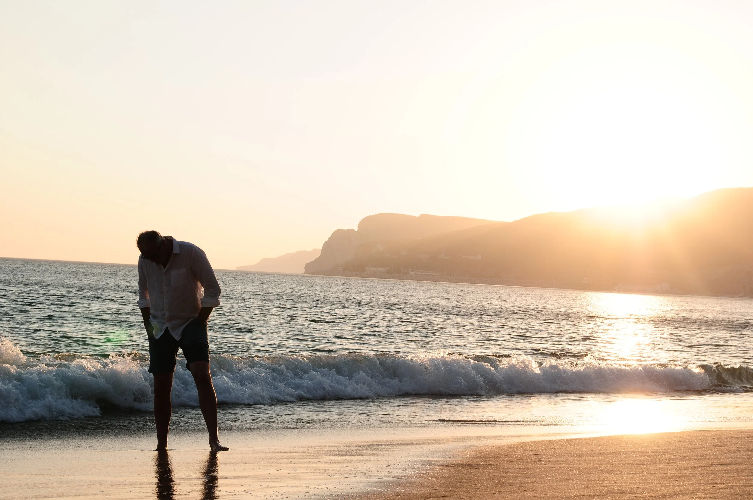 Silhouette of a man walking along the beach during sunset, with ocean waves and a hilly coastline in the background.