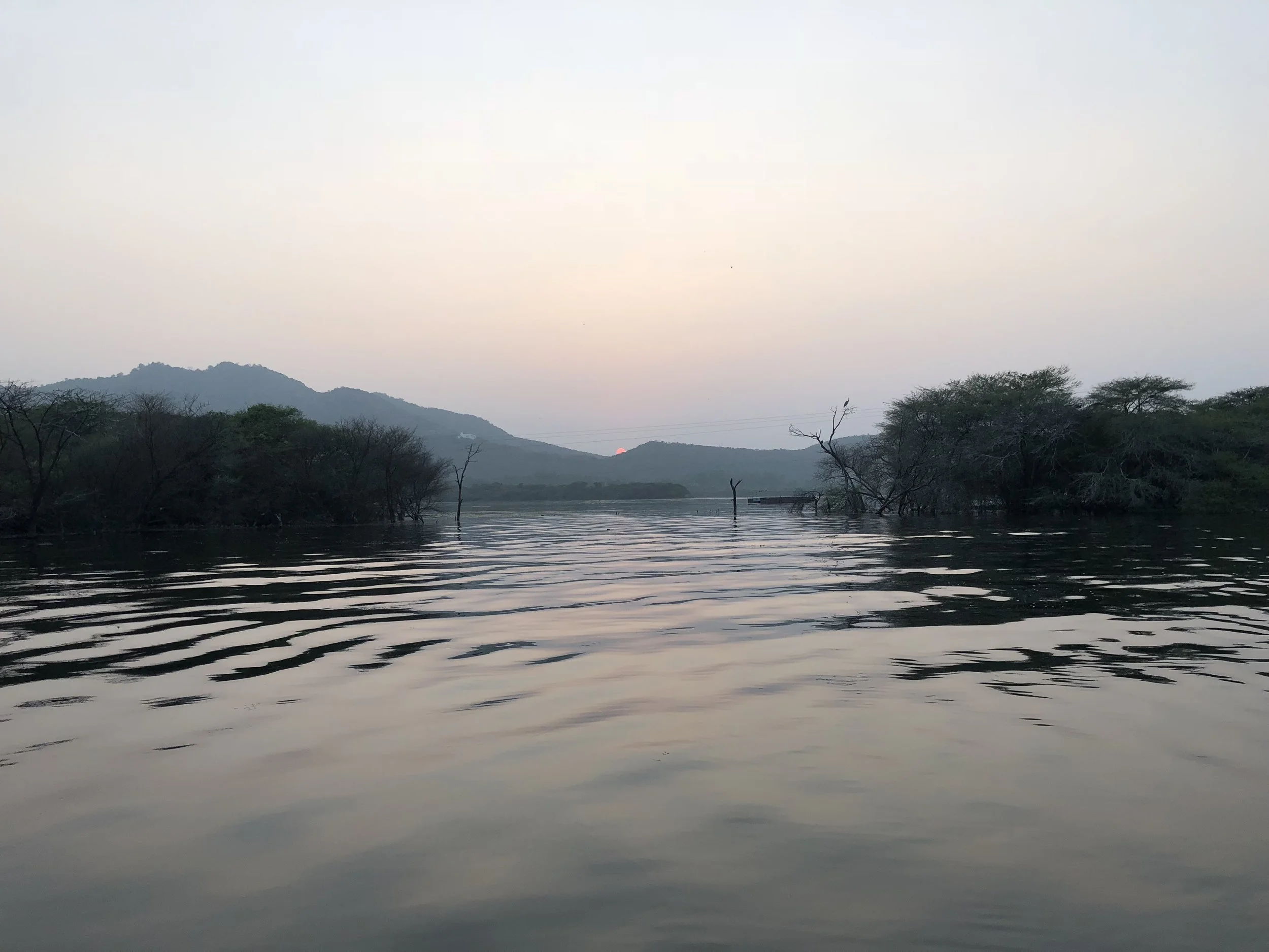 A serene river scene at sunset or sunrise, with rippling water, distant hills, and sparse trees along the riverbank, some partially submerged.