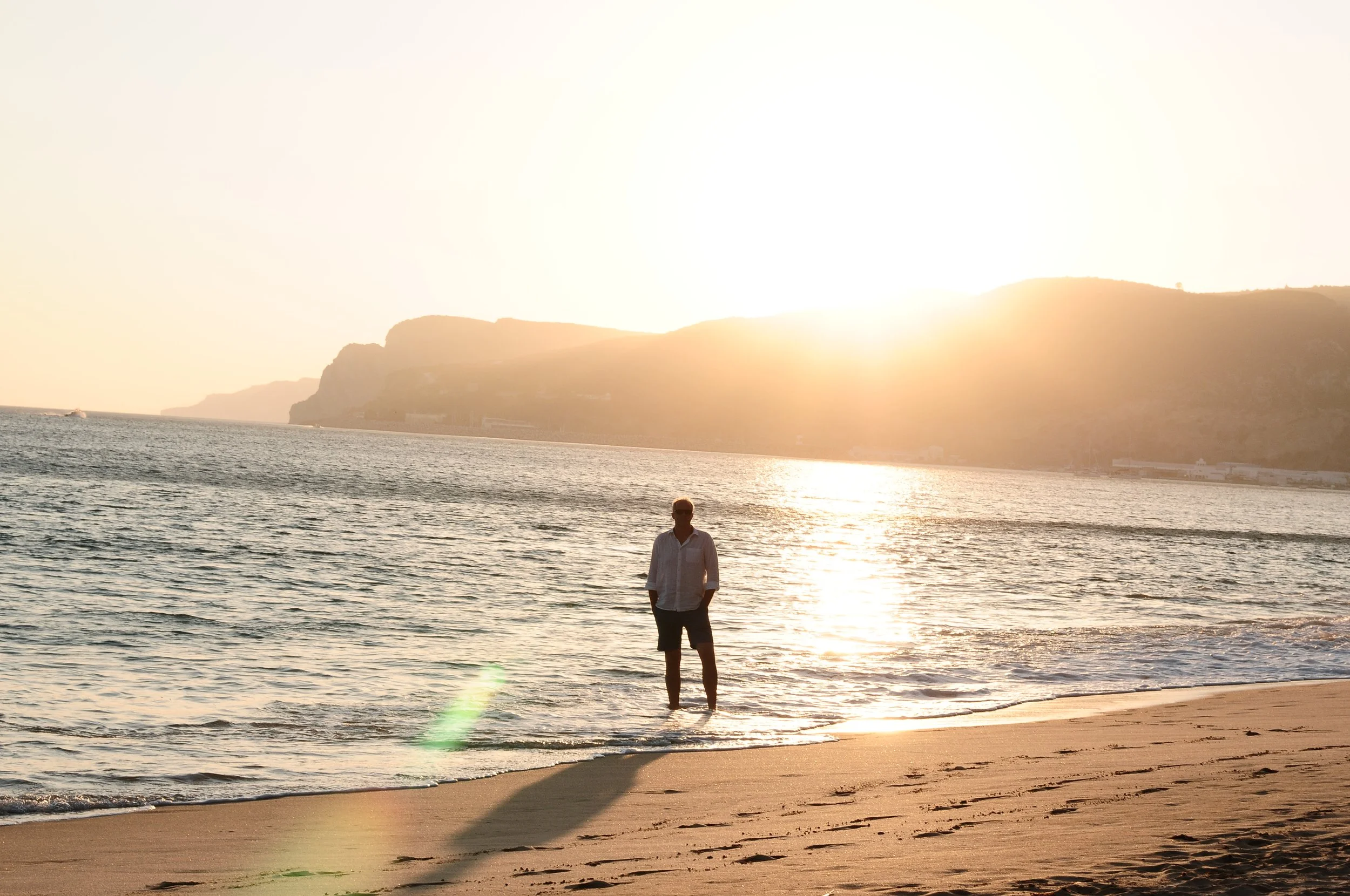 Person standing on a sandy beach near the water at sunset, with hills and cliffs in the background.
