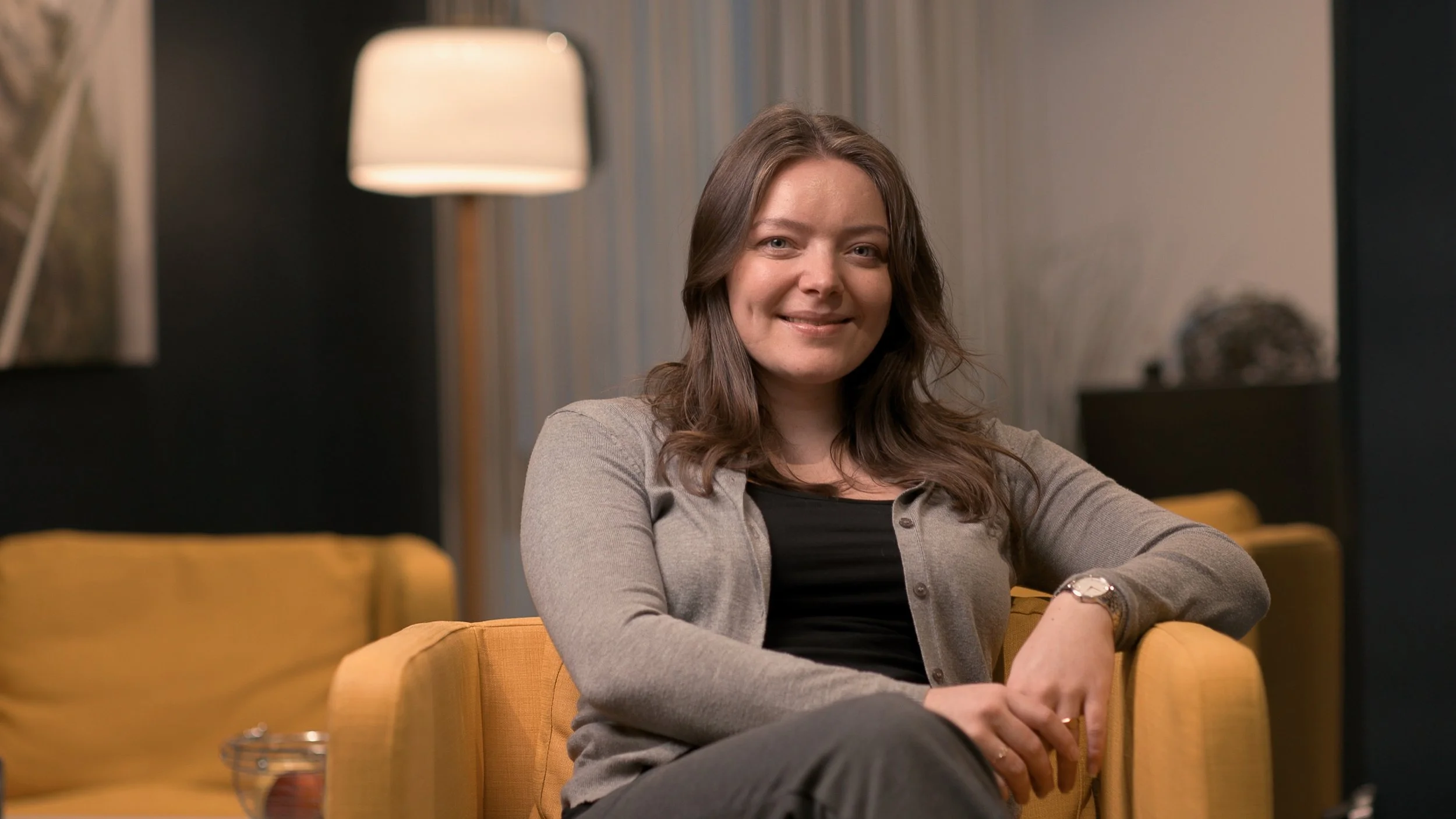 Kvinne med brunt, krøllet hår sitter i en gul sofa i et rom med moderne innredning, smilende og ser mot kameraet.