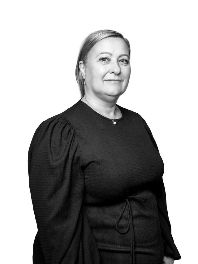 En svart-hvitt portrett av en kvinne som står foran en lys bakgrunn, iført svart kjole med ballongermer og en liten perlesmykke.