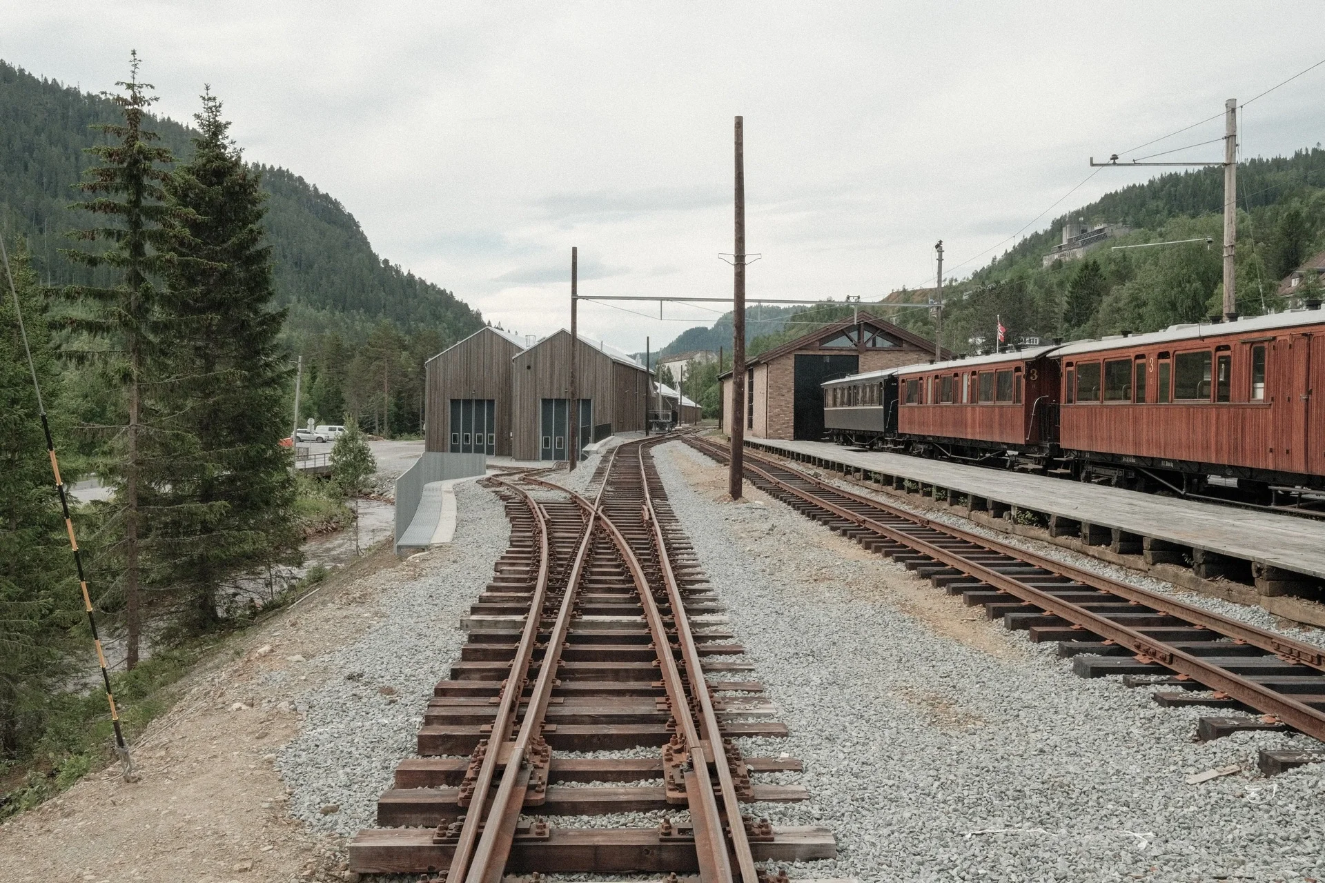 Jernbanelinje med spor og bygninger i fjellandskap, omgitt av trær og fjell.