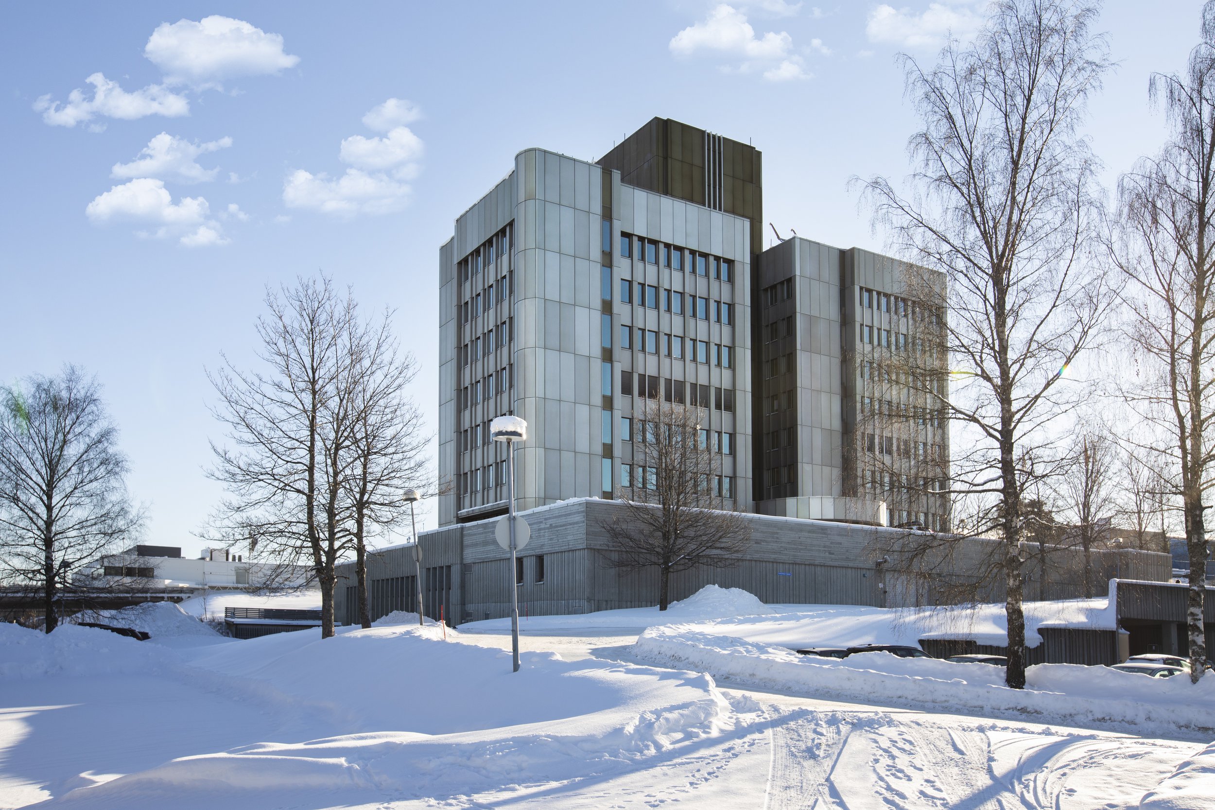 Snødekt bygningskompleks omgitt av flere nakne trær under en klar blå himmel.