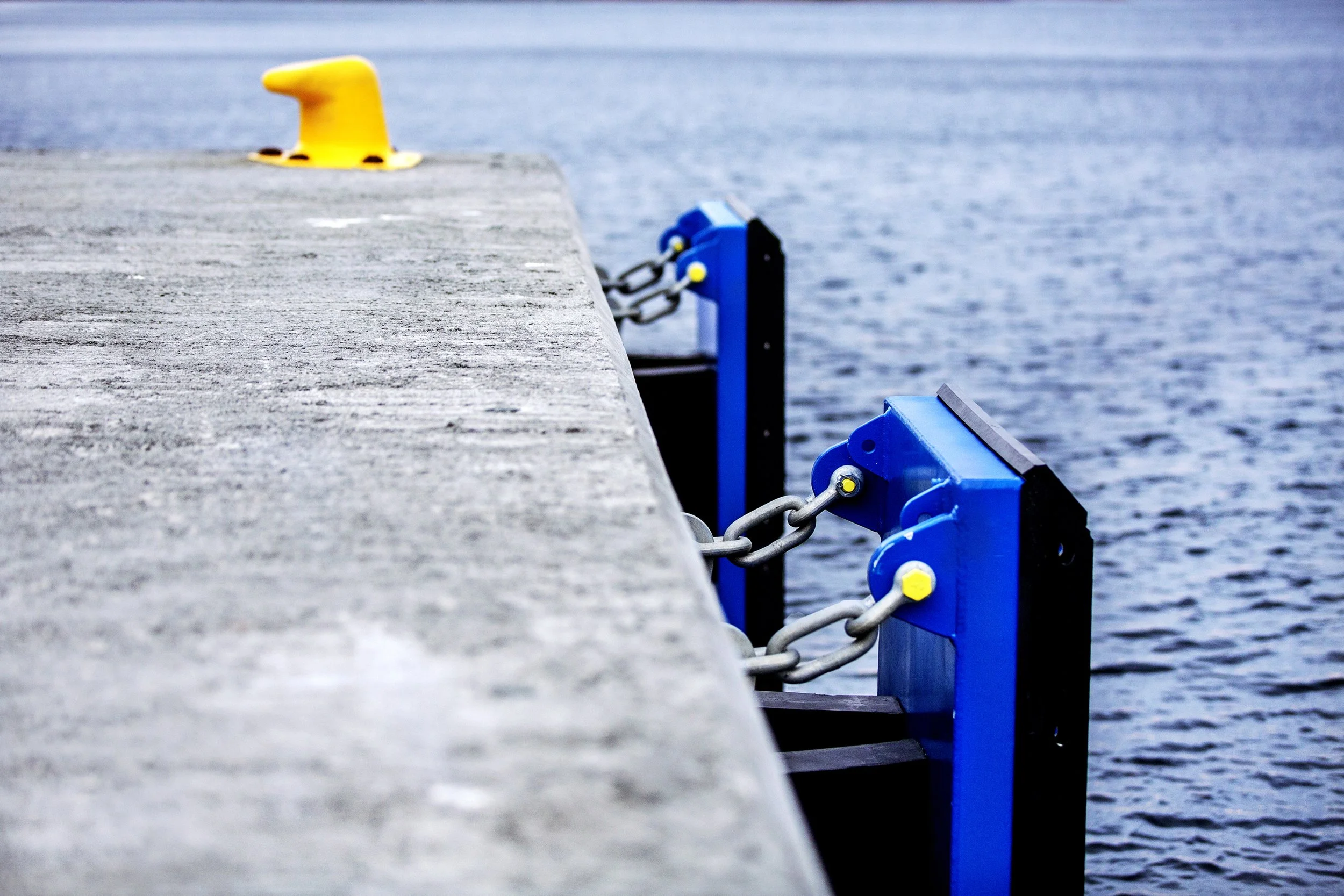 Bild av en brygge med blå og gul rykkere og tau, ut mot en vannfylt bakgrunn.