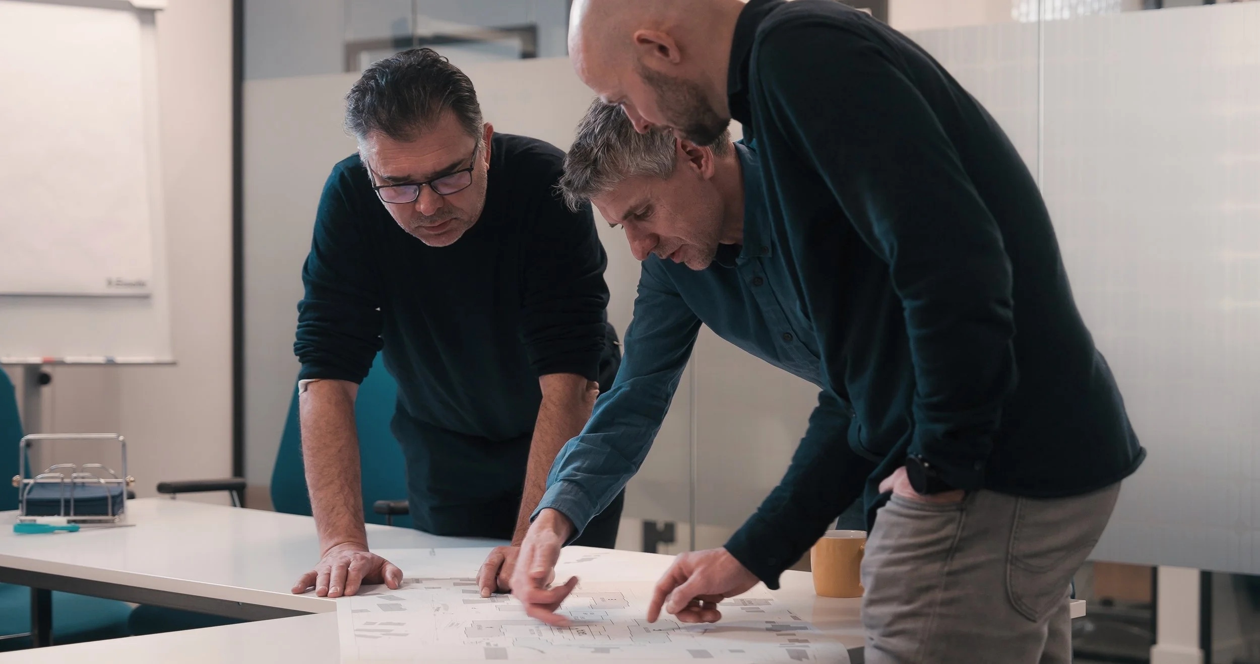 Tre menn i arbeidsantrekk diskuterer et ark med planer på et bord i et kontormiljø.