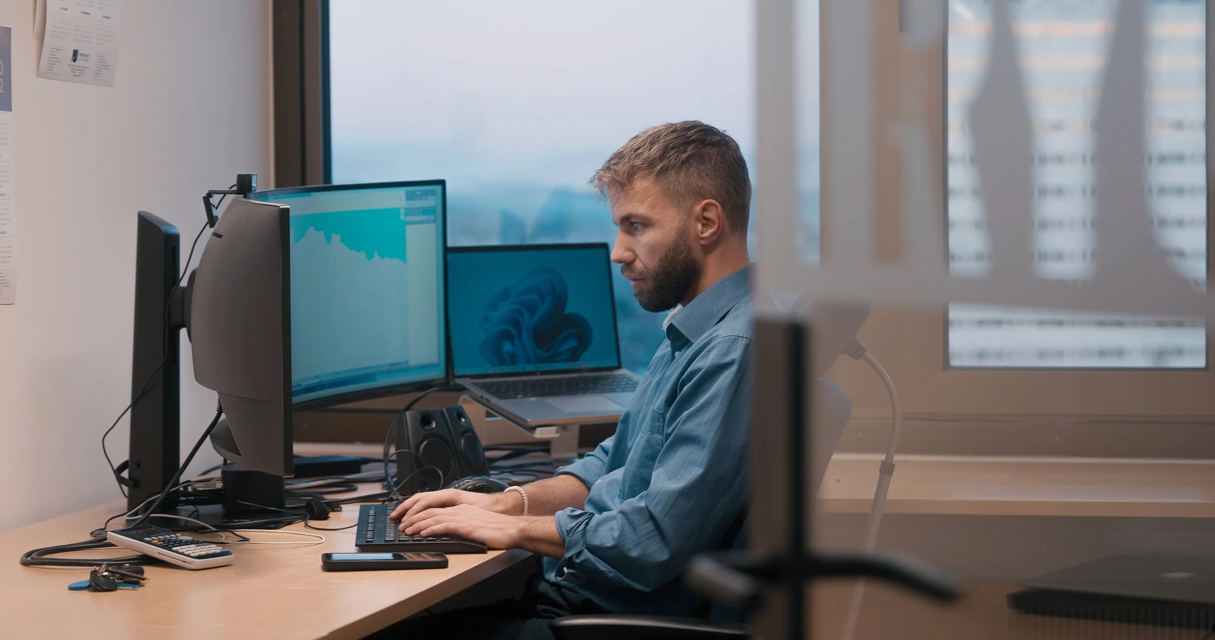 En mann jobber ved et kontorbord med to skjermer, en bult med laptop, en kalkulator og nøkkelknippe.
