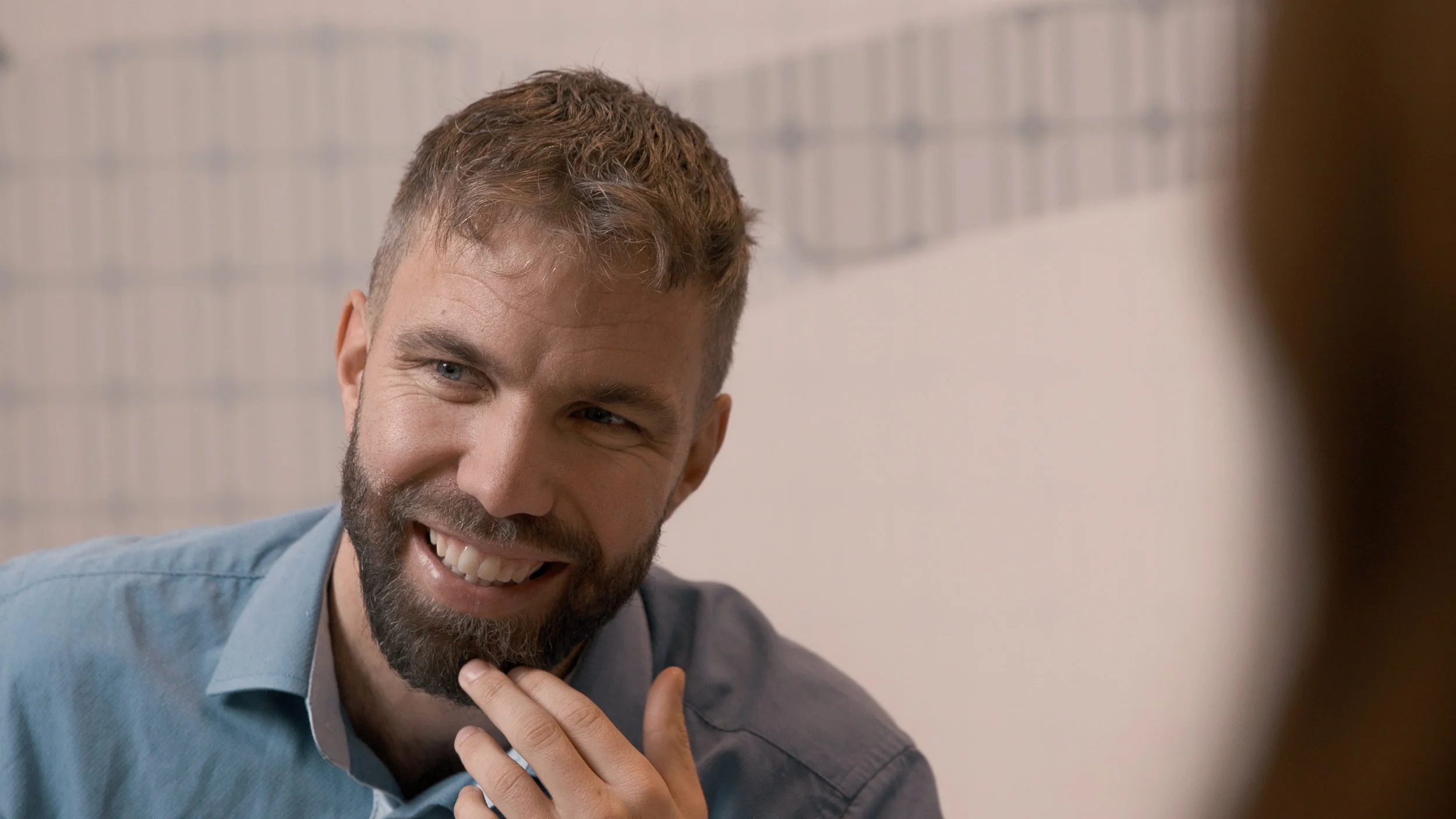 En mann med skjegg og kort, brunt hår smiler mens han holder hånden mot hake. Han er iført en lys blå skjorte og sitter ved en vegg med lyse murstein.