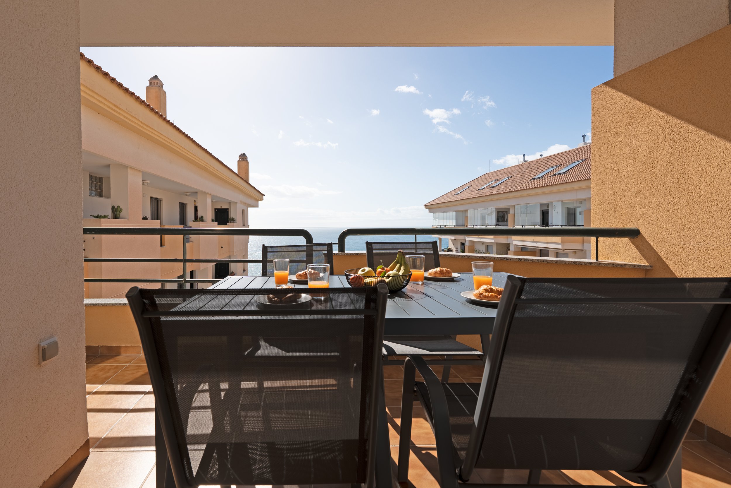 Breakfast on a balcony with a view of neighboring buildings, ocean in the distance, and a partly cloudy sky.