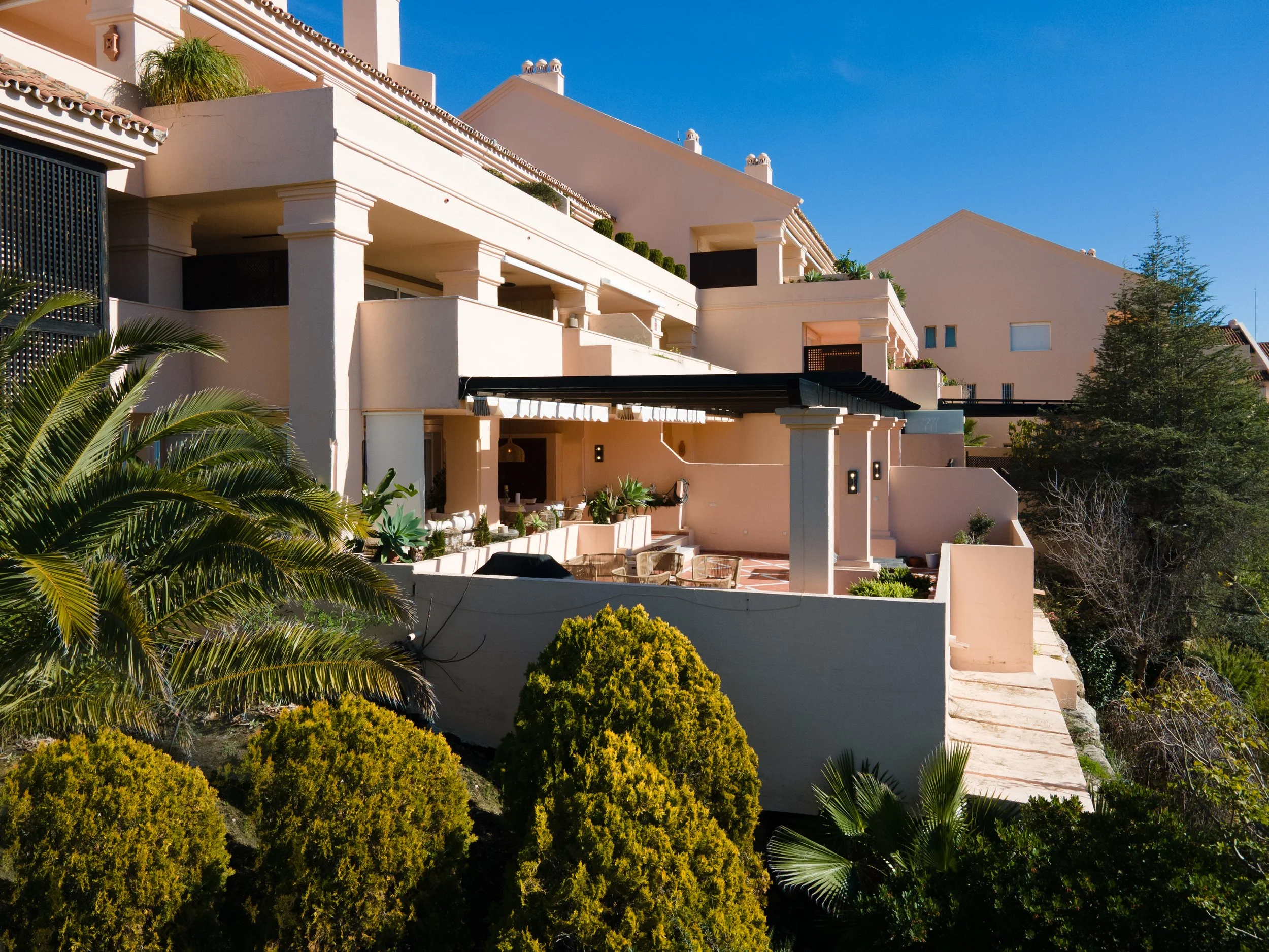 Multi-story modern house with white exterior, balconies, outdoor seating area, and lush greenery under a blue sky.
