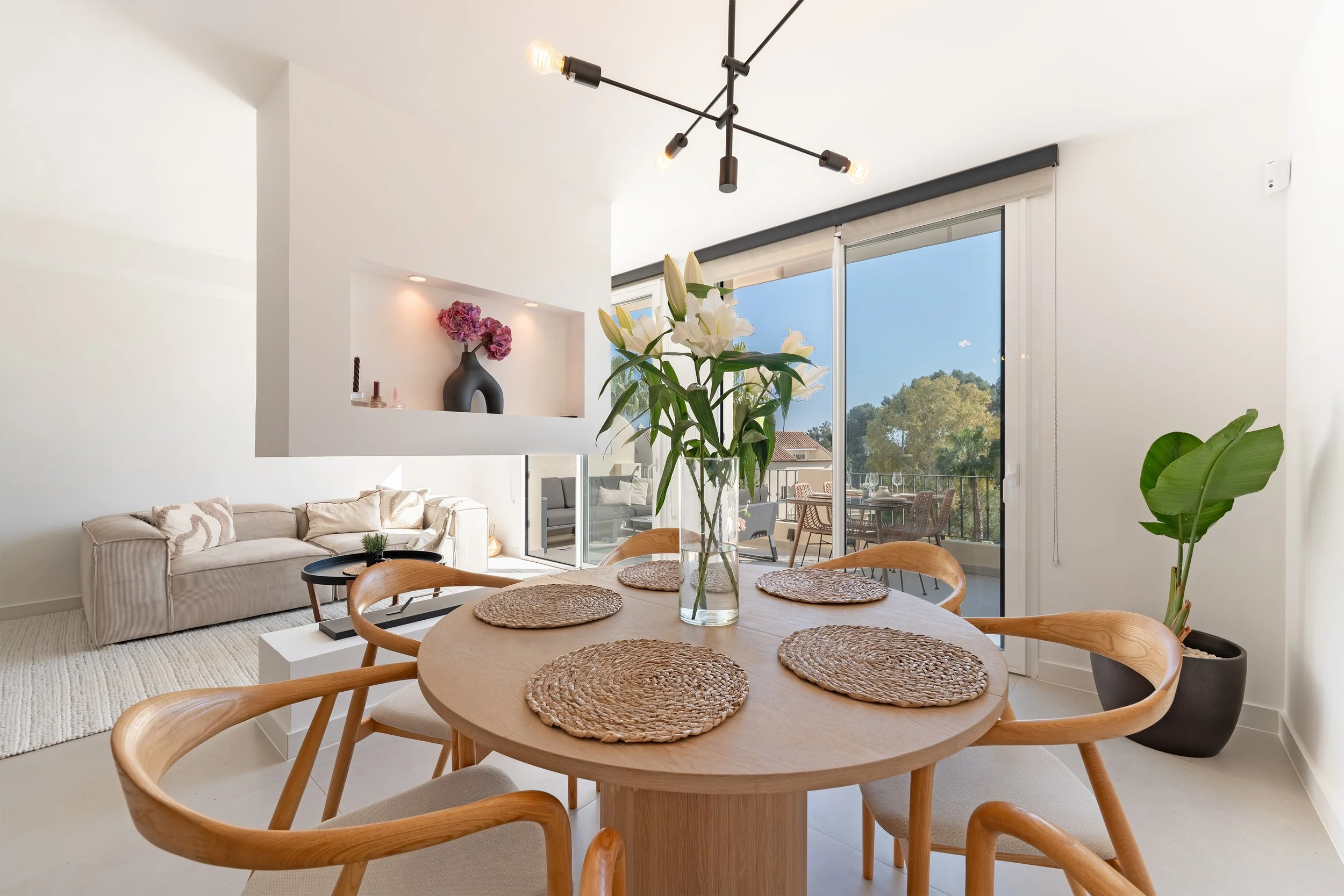 Dining area with a round wooden table, six woven placemats, a glass vase with white lilies, and wooden chairs. Behind, there is a living room with a beige sofa, a small black side table, and a built-in wall shelf with a black vase and pink flowers. L