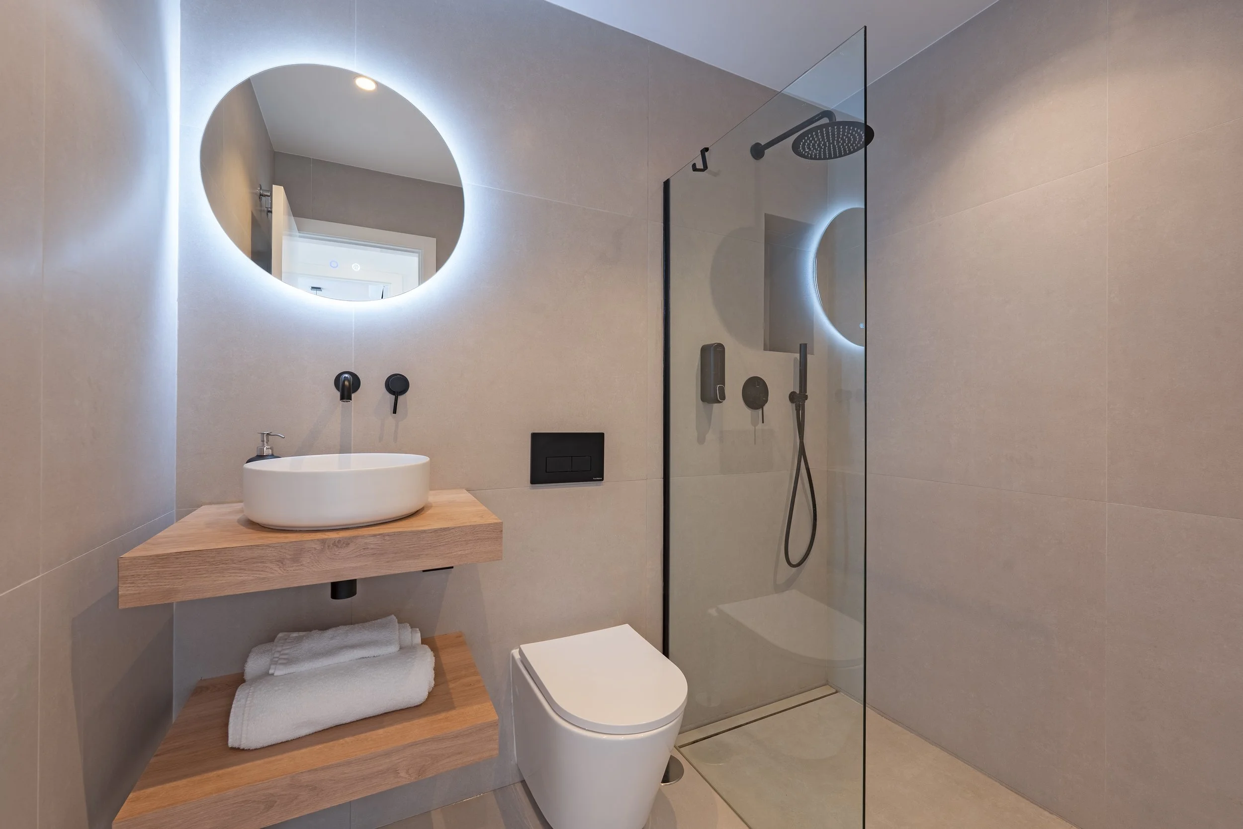 Modern bathroom with beige tiled walls, a round backlit mirror, a wall-mounted sink with a wooden shelf underneath, white towels, a wall-mounted toilet, and a walk-in shower with a glass divider and black fixtures.