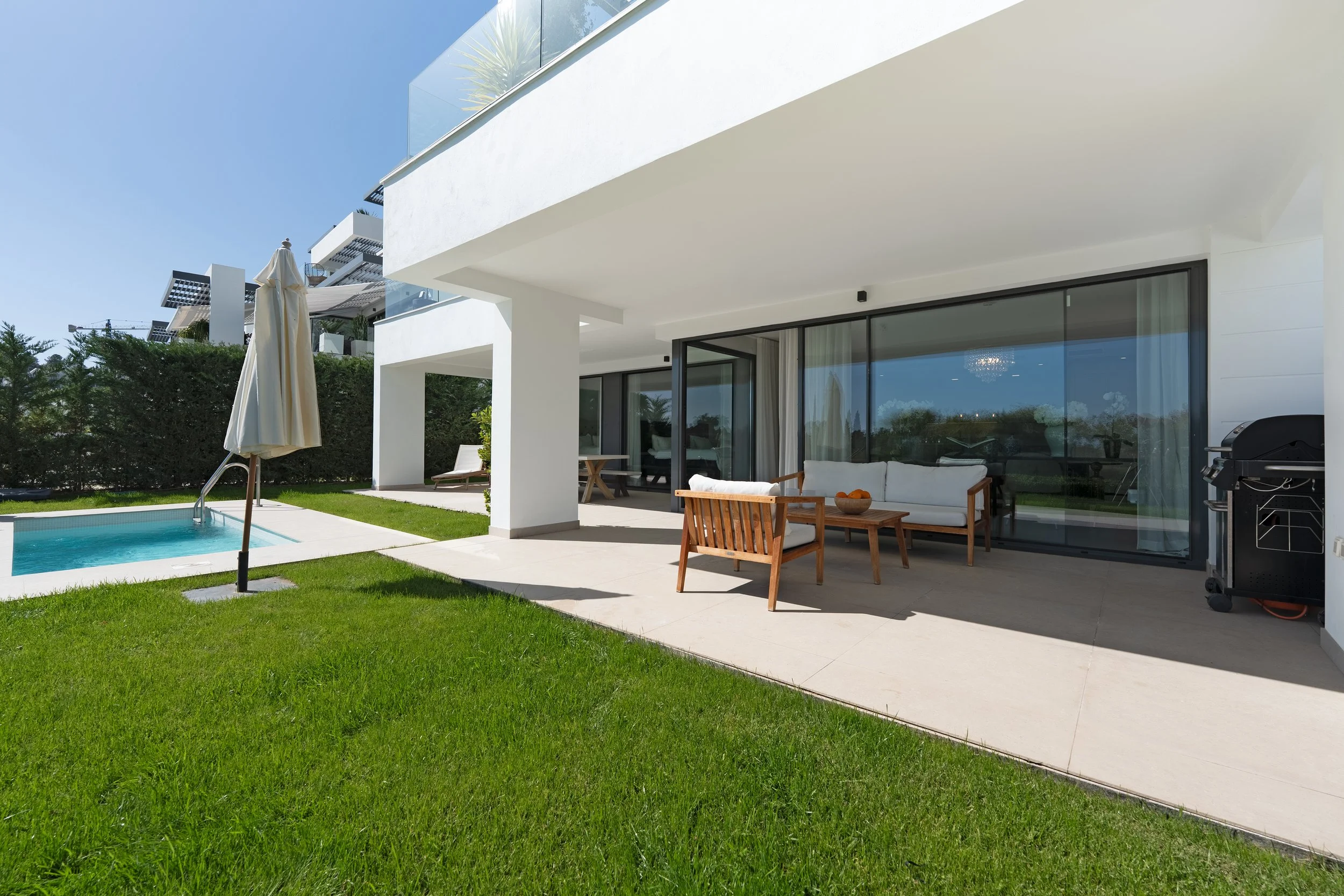 Modern house exterior with patio, outdoor seating, umbrella, small pool, and green lawn under a clear blue sky.