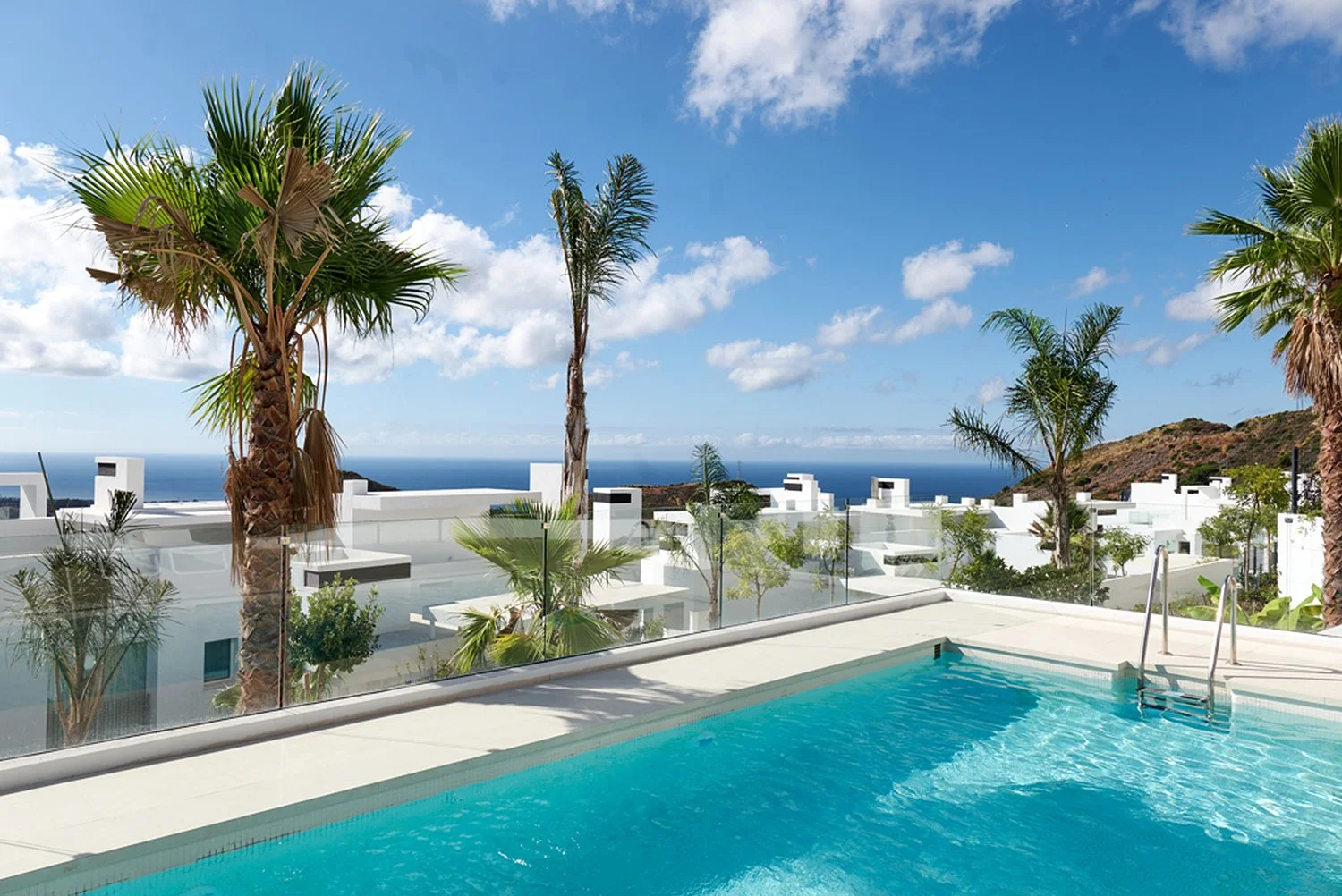 A rooftop swimming pool with a view of a blue ocean, surrounded by palm trees and white modern buildings under a partly cloudy sky.