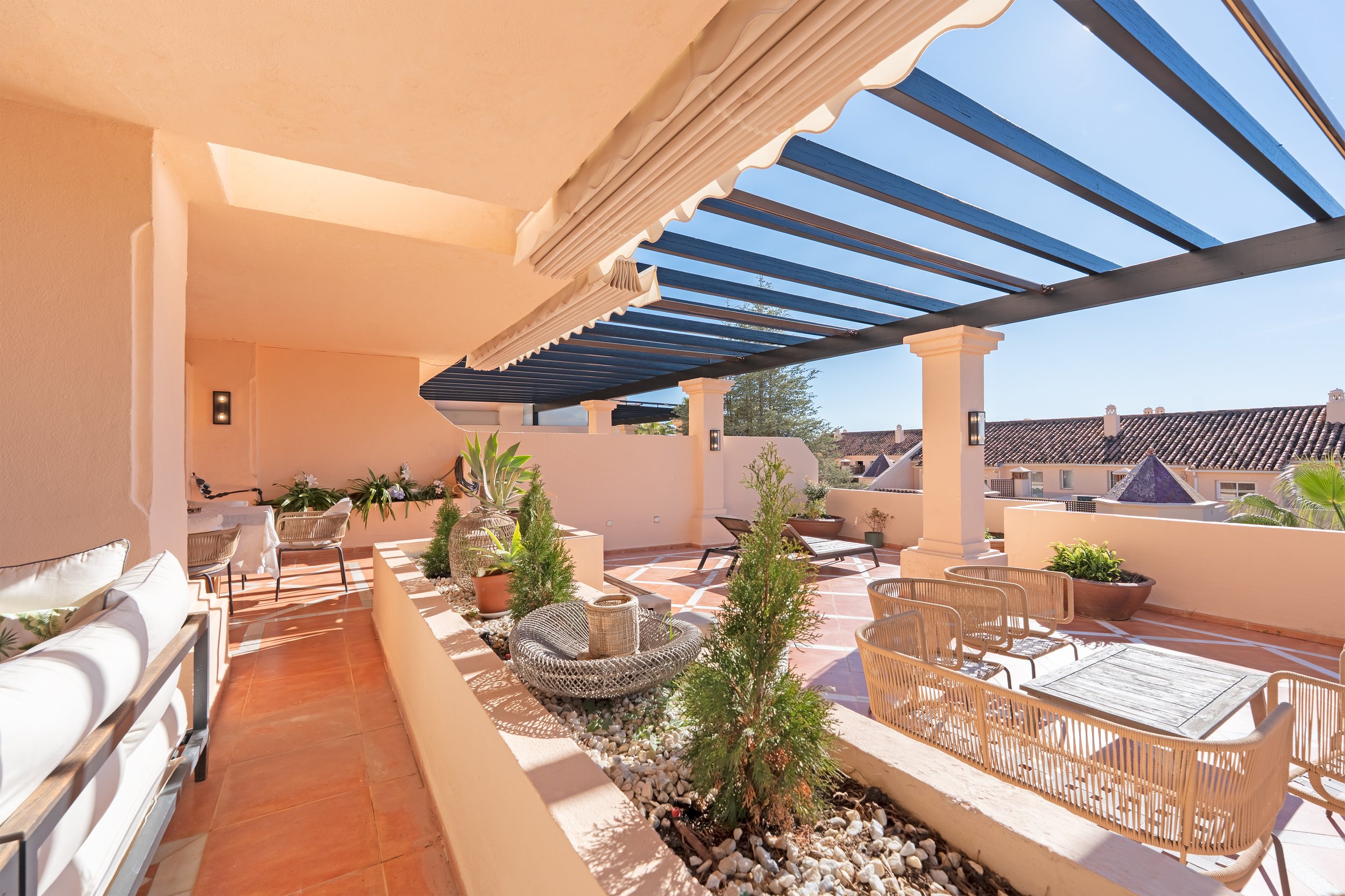 Sunny outdoor terrace with potted plants, lounge chairs, woven chairs, a table, and a blue pergola.