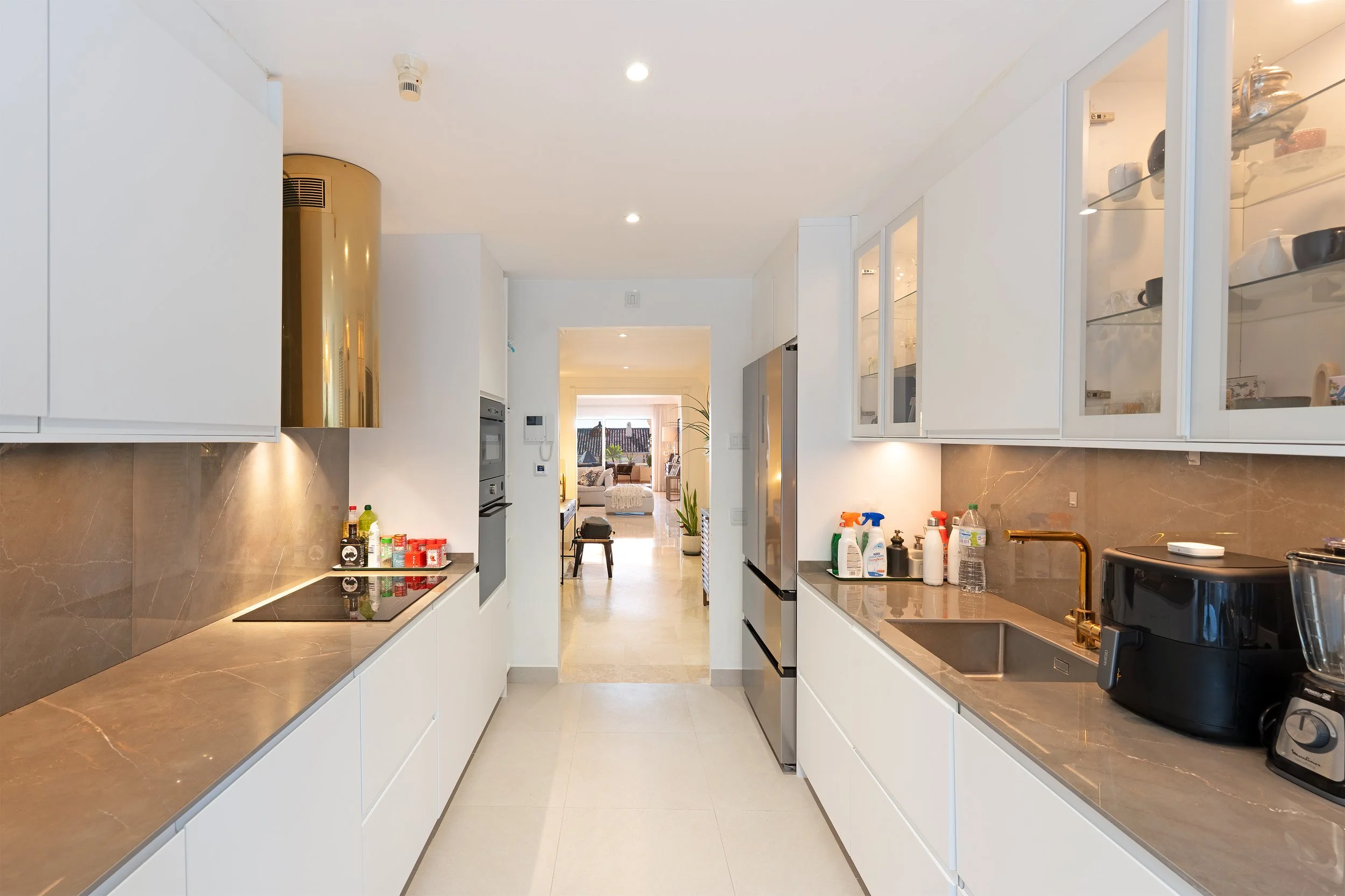 Modern kitchen with white cabinets, brown marble countertops, and stainless steel appliances. Visible are cleaning supplies, a black air fryer, and a small kitchen sink. Beyond the kitchen, a living room with large windows and seating area is visible