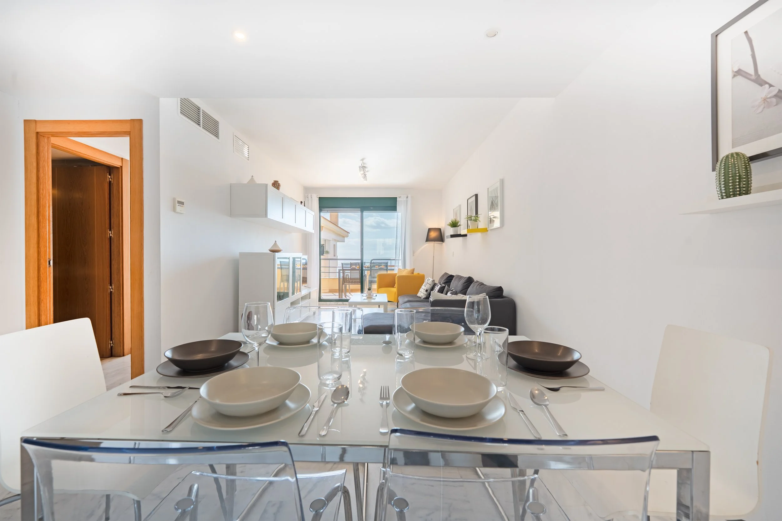 Modern dining room and living room with a glass table, white chairs, and a gray sofa with a yellow armchair, overlooking a balcony with a view and sliding glass door.