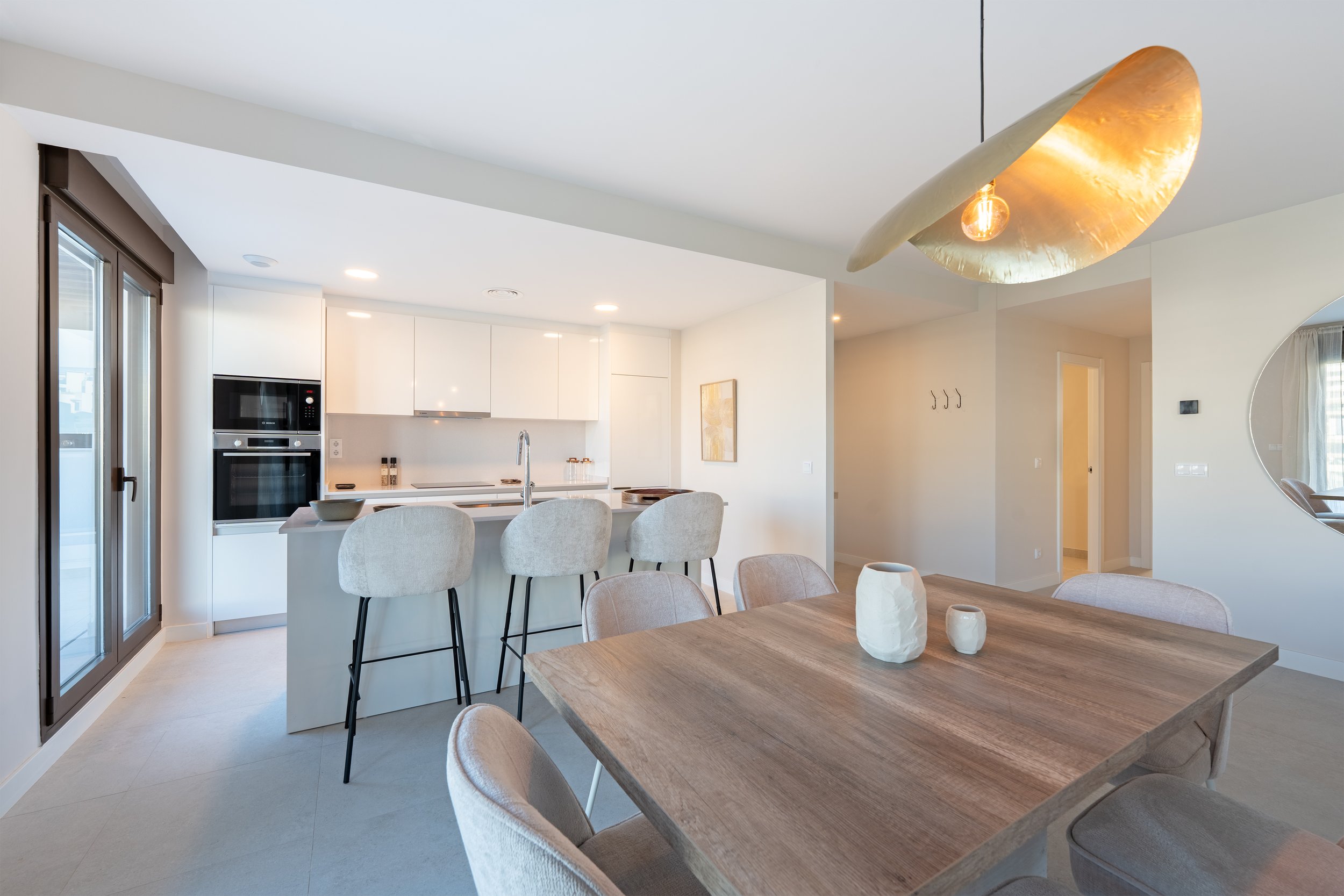 Modern open-concept kitchen and dining area with white cabinetry, a wooden dining table, light-colored chairs, and a unique gold-colored ceiling light fixture.
