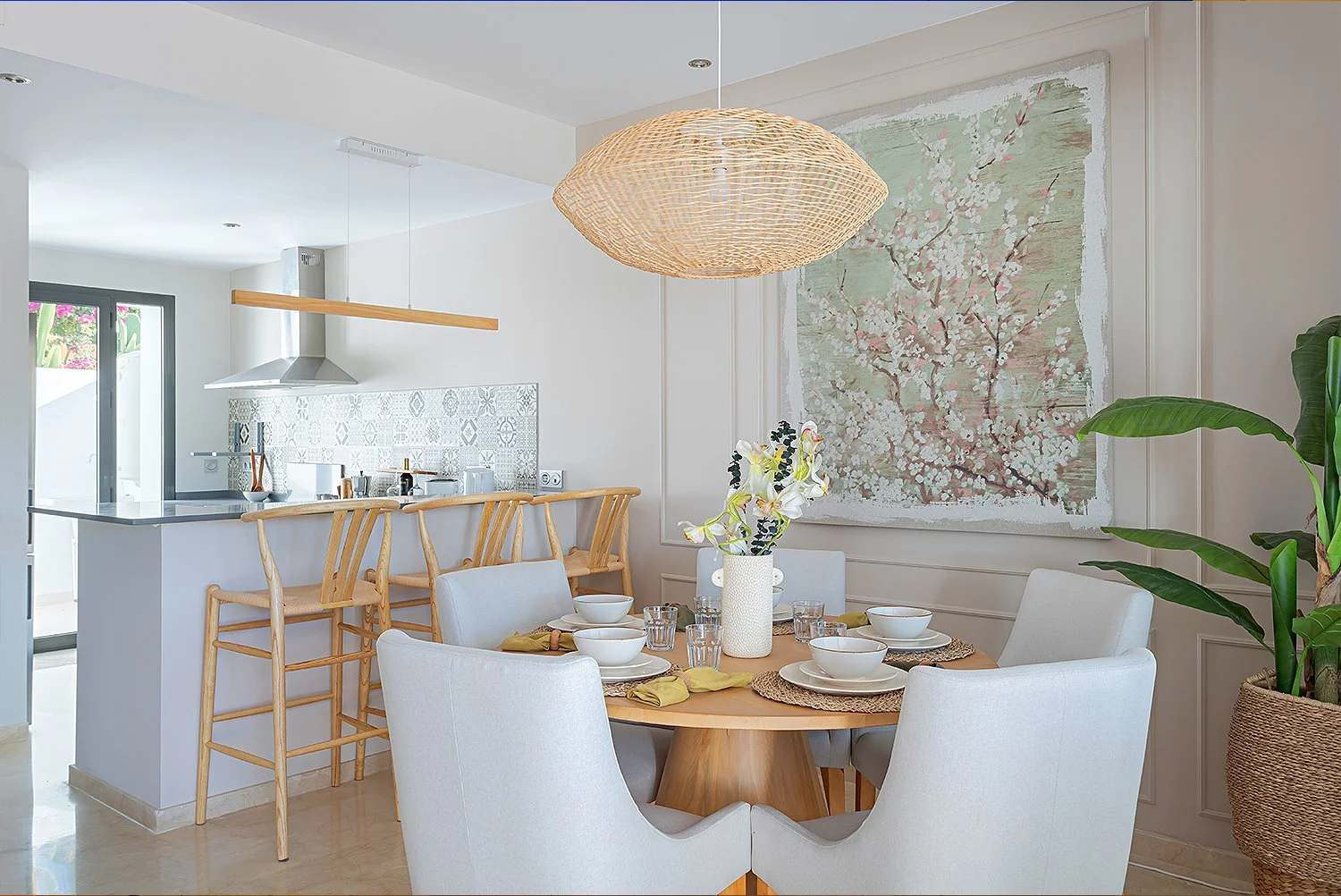 A dining area and kitchen with a round wooden table set with white bowls, glasses, and yellow napkins, surrounded by white upholstered chairs. A large woven pendant light hangs above the table. Behind the table is a large textured painting of blossom