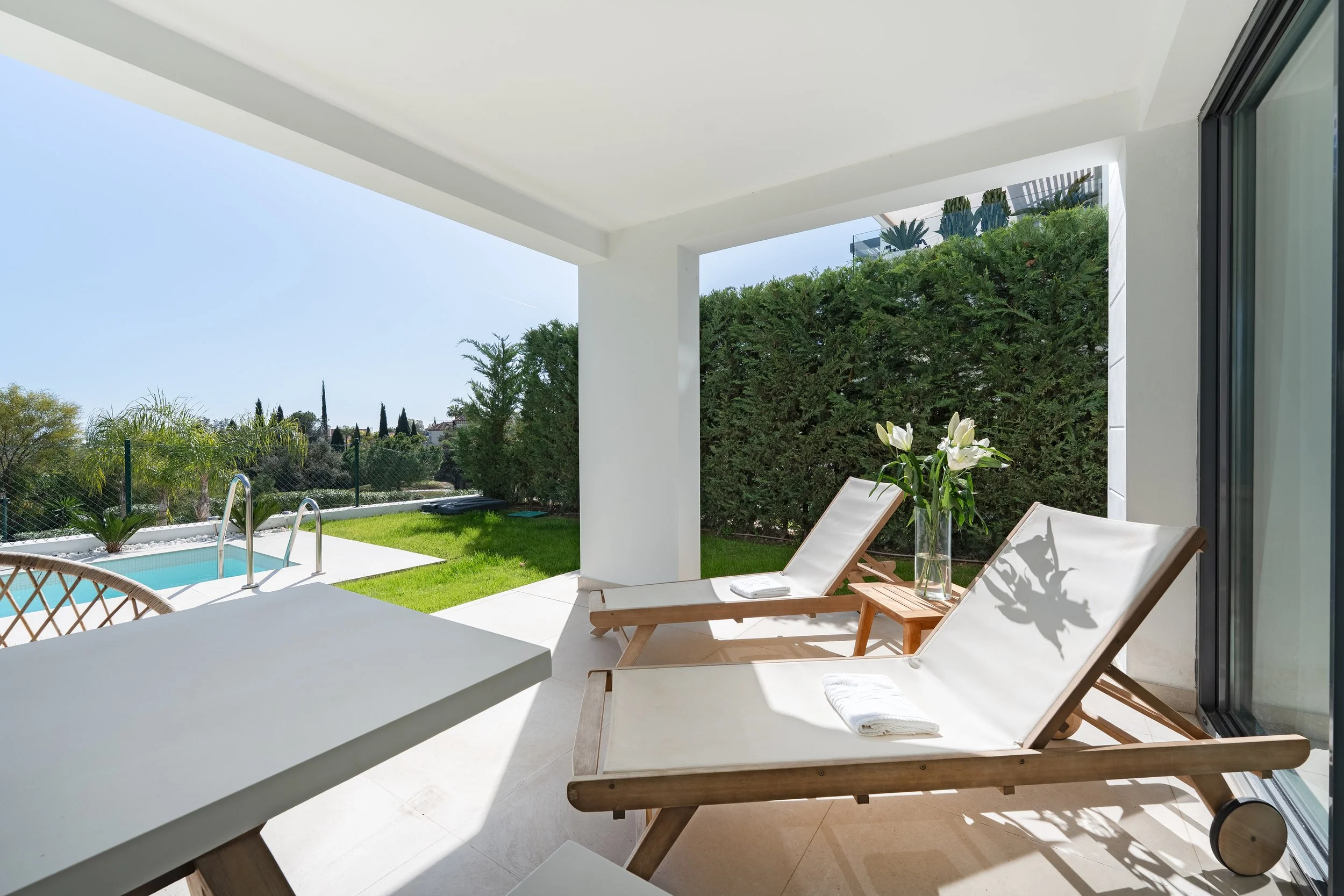 Outdoor patio area with two wooden lounge chairs, white cushions, towels, a small wooden table with a vase of white lilies, a small swimming pool, green lawn, and trees in the background under a clear blue sky.