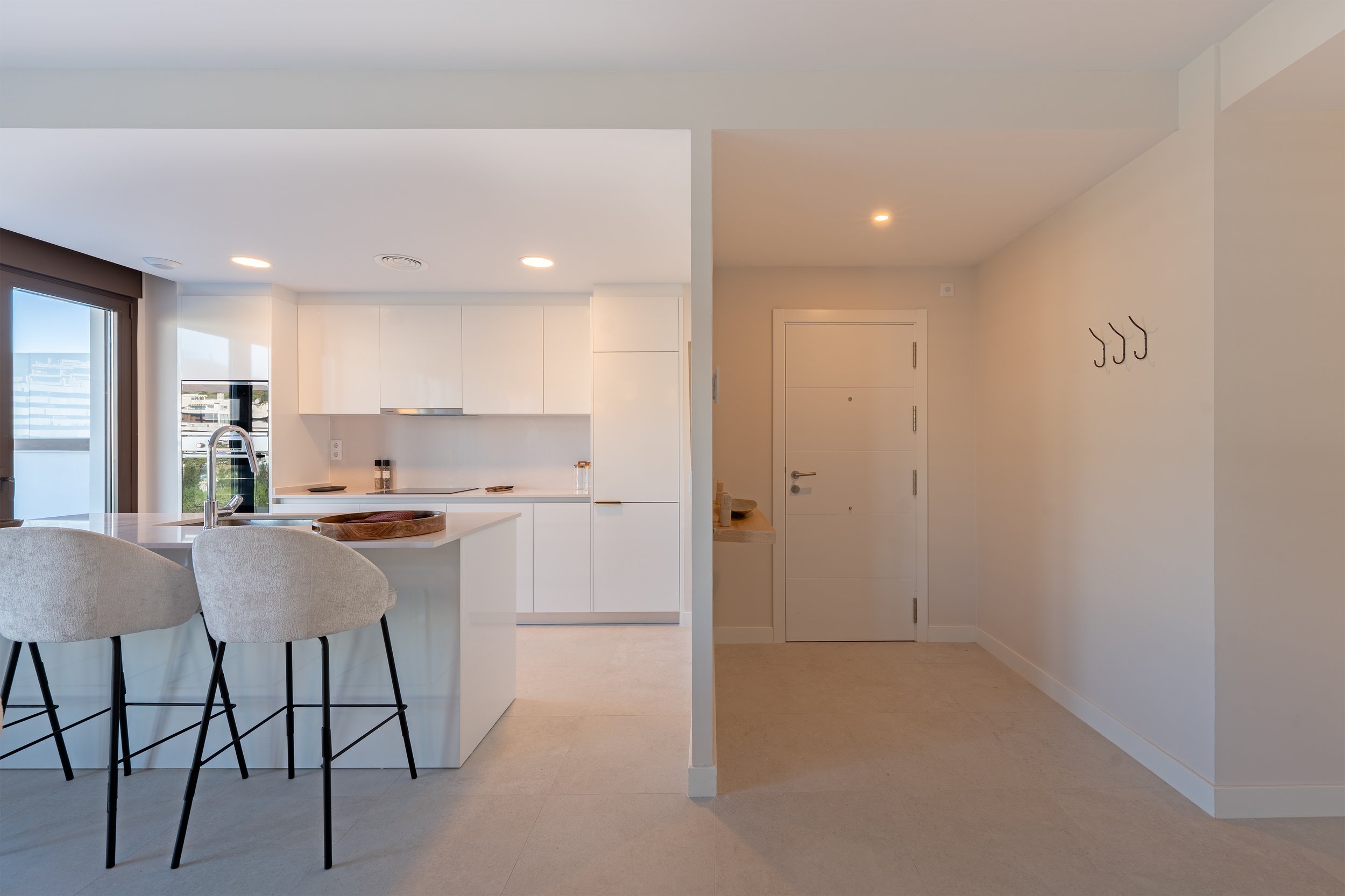 Modern kitchen with white cabinets, a breakfast bar with two beige upholstered stools, a large window, and a city view, adjacent to an entryway with hooks on the wall.