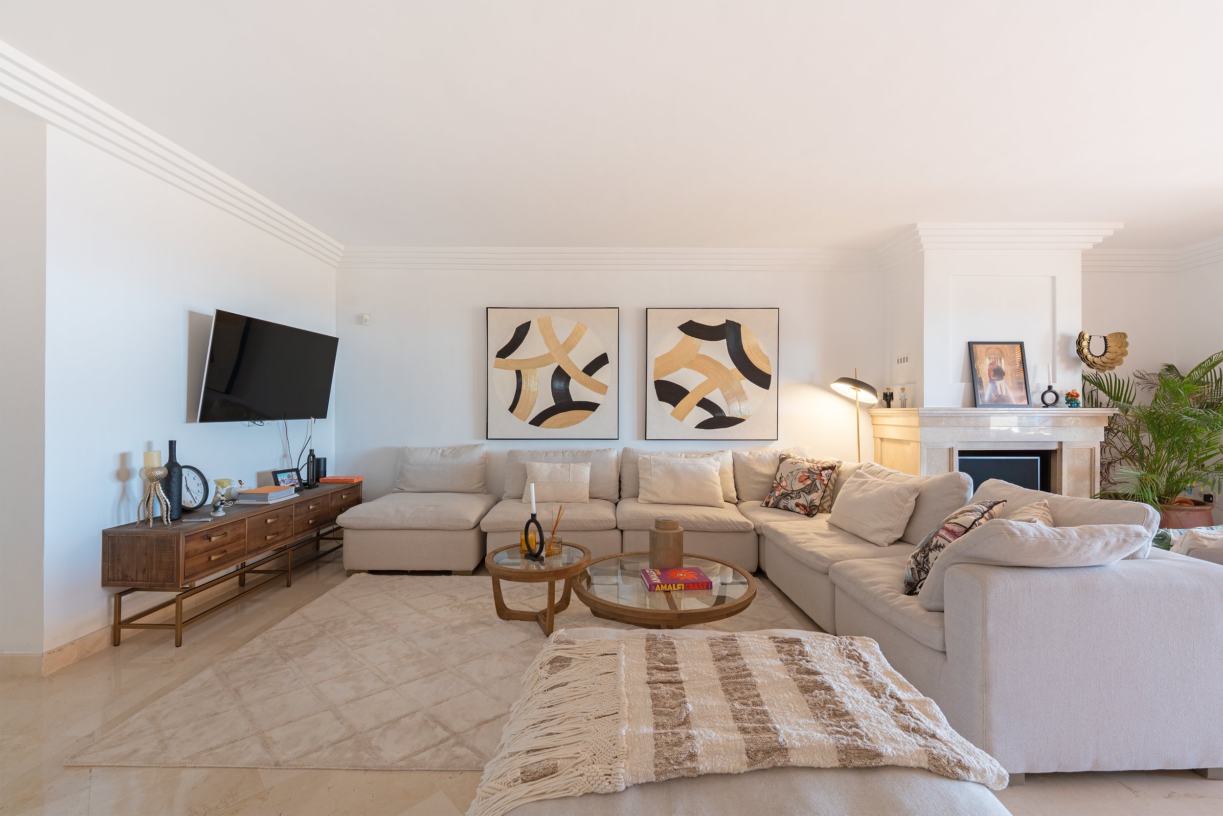 Living room with white sectional sofa, two round coffee tables, a wall-mounted television, framed abstract art, fireplace, decorative pillows, and houseplants.