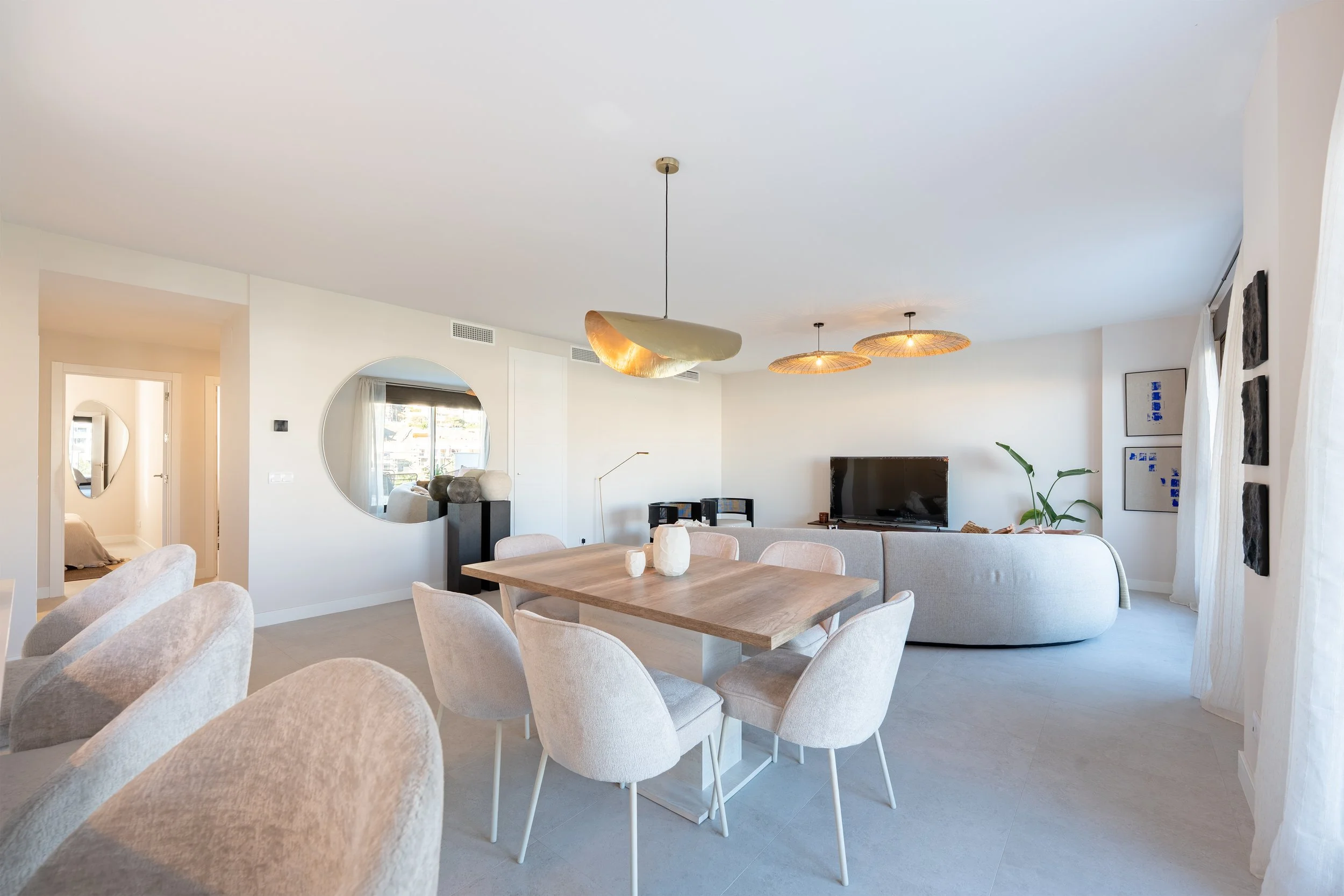 Modern living and dining room with white walls, light-colored chairs, a wooden dining table with decorative vases, a gray curved sofa, a flat-screen TV, and wall art, illuminated by natural light through large windows with white curtains.