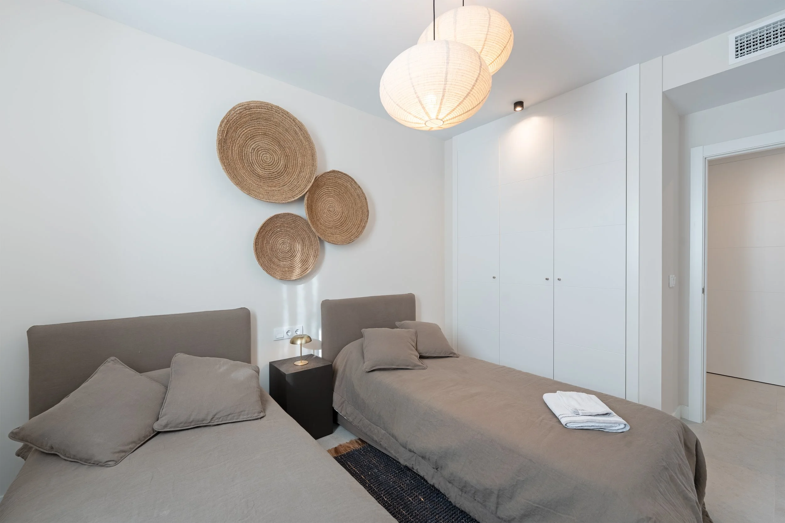 A modern bedroom with two beds, beige bedding, and pillows, a black nightstand with a small lamp, white walls, woven wall art, and a large white wardrobe.