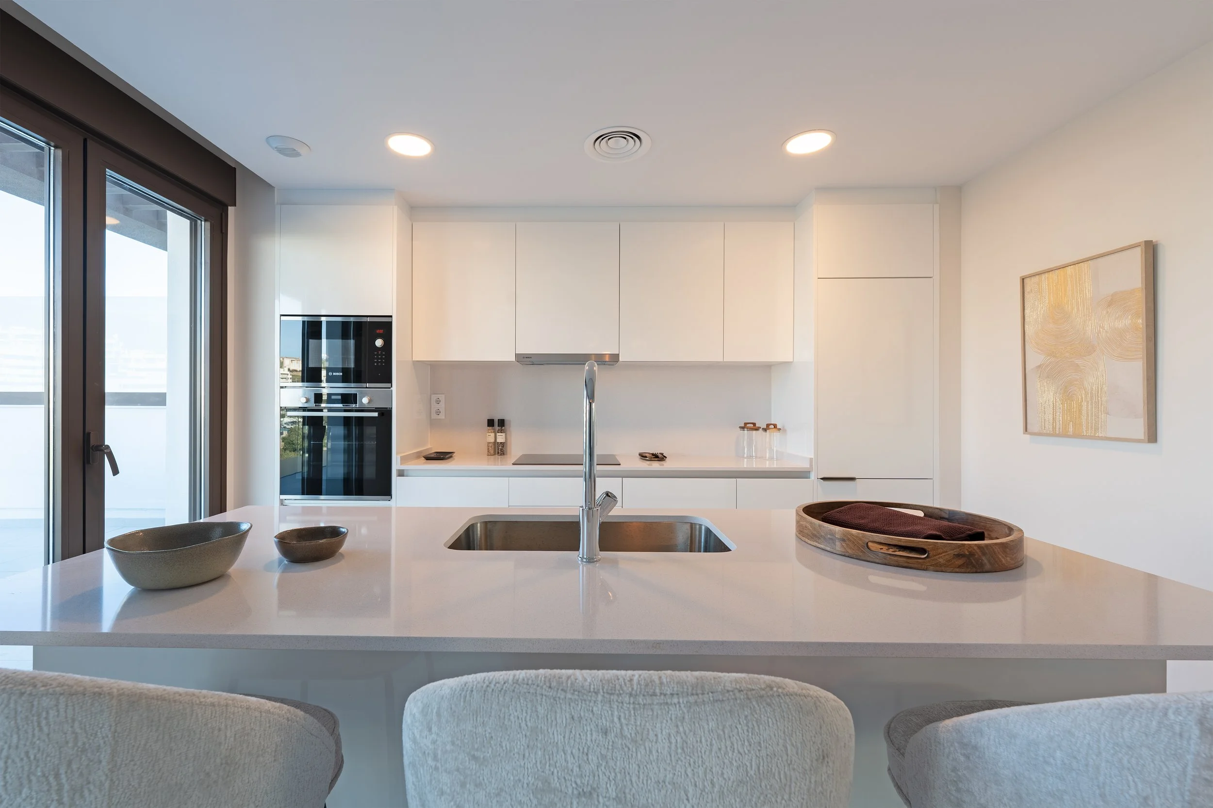 Modern kitchen with white cabinets, island with a sink, and beige chairs, lit by recessed ceiling lights, with artwork on the wall and sliding glass door.