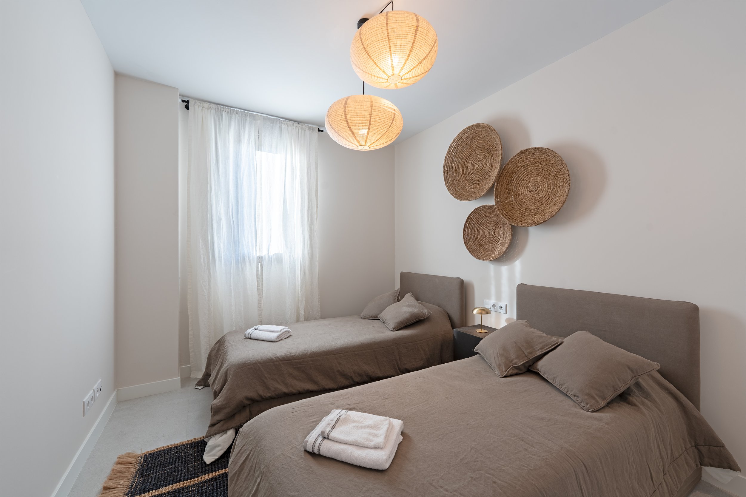 A bedroom with two beds, beige bedding, and matching pillows, a small black nightstand with a gold lamp, a wall with three decorative woven baskets, a window with sheer white curtains, and a ceiling with two round paper lantern light fixtures.