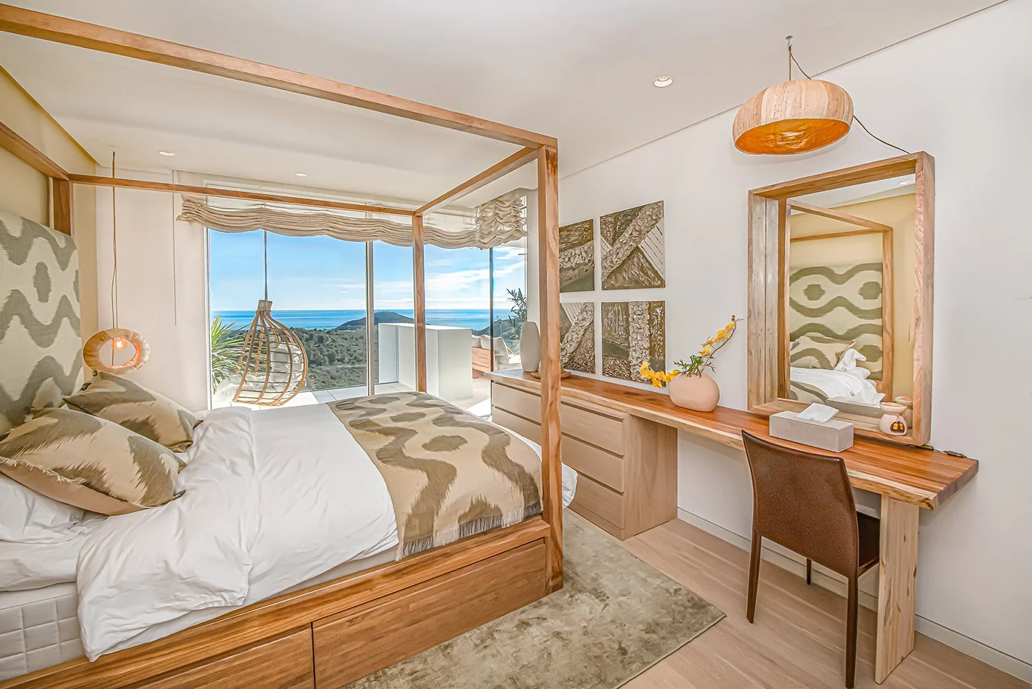 Modern bedroom with a sliding glass door overlooking a view of the ocean and mountains, featuring a wooden four-poster bed with patterned bedding, a hanging wicker chair, a wooden dresser, and a wooden desk with a chair and a large mirror.
