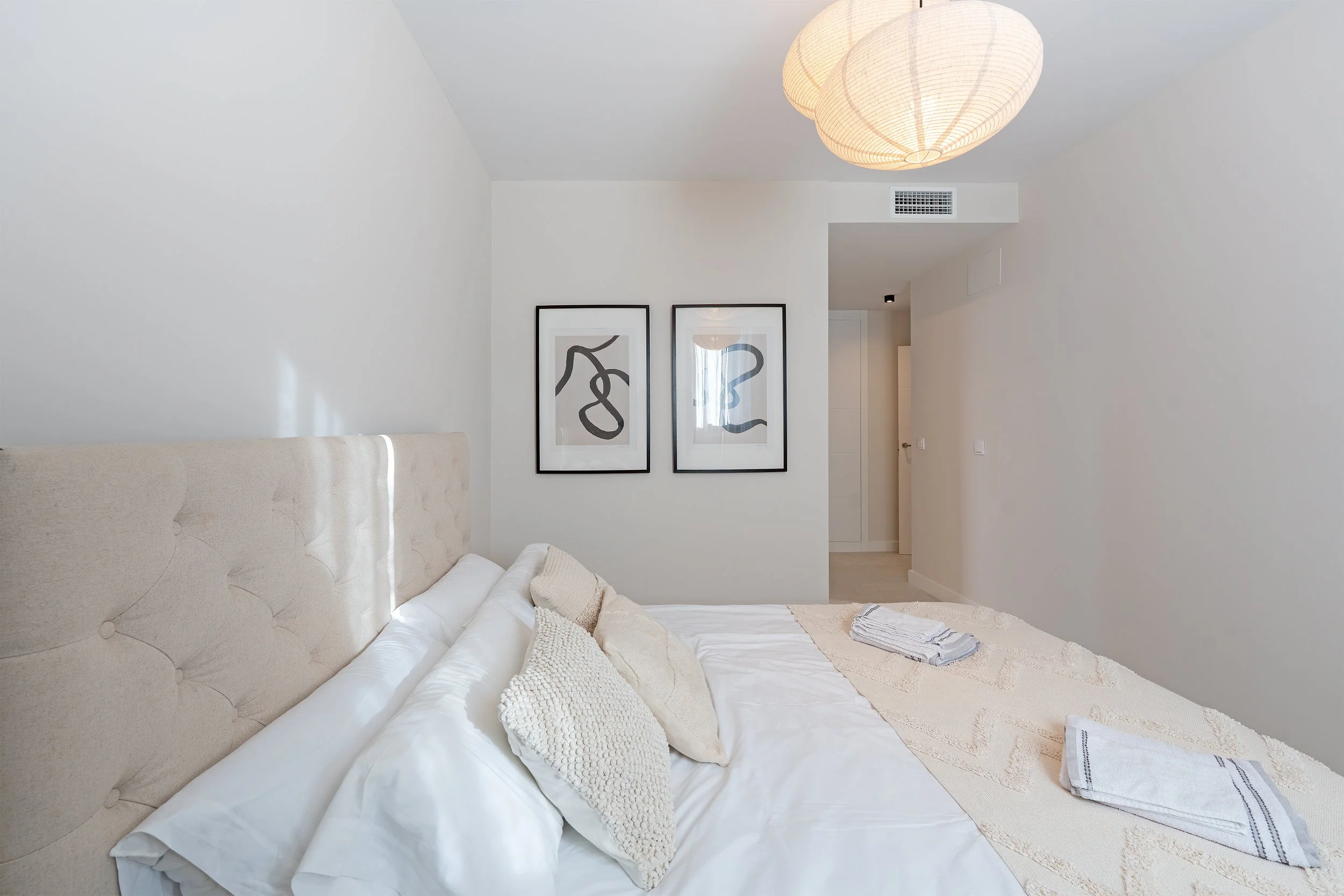 A neatly made bed with white linens and beige pillows in a minimalist white bedroom. Two abstract black and white framed artworks hang on the wall above, and a modern paper lantern hangs from the ceiling. Towels are folded on the bed.