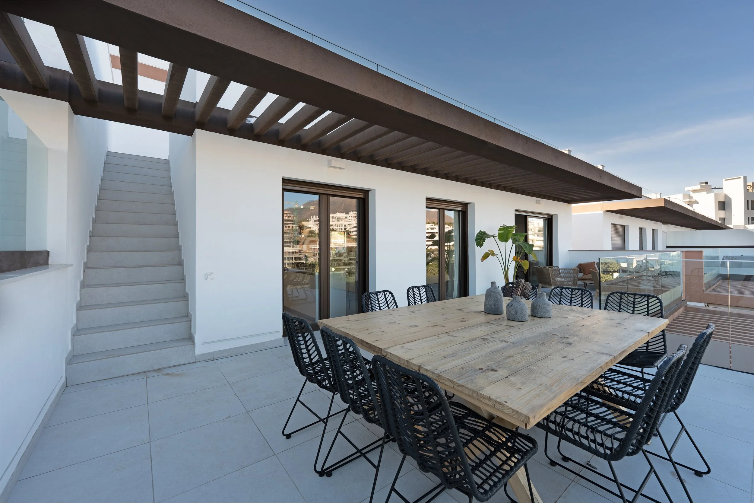 Outdoor rooftop patio with a wooden dining table, black chairs, potted plants, and cushioned seating area, with city buildings in the background.