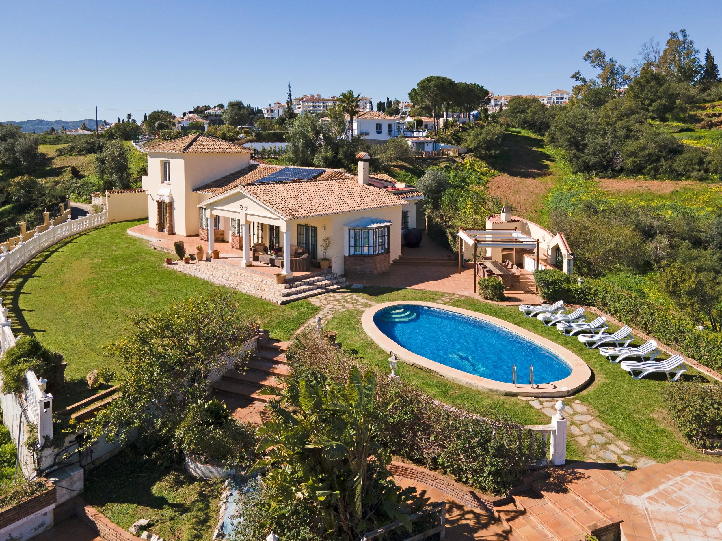 A house with a swimming pool in the backyard, surrounded by greenery and lounge chairs, under a clear blue sky.