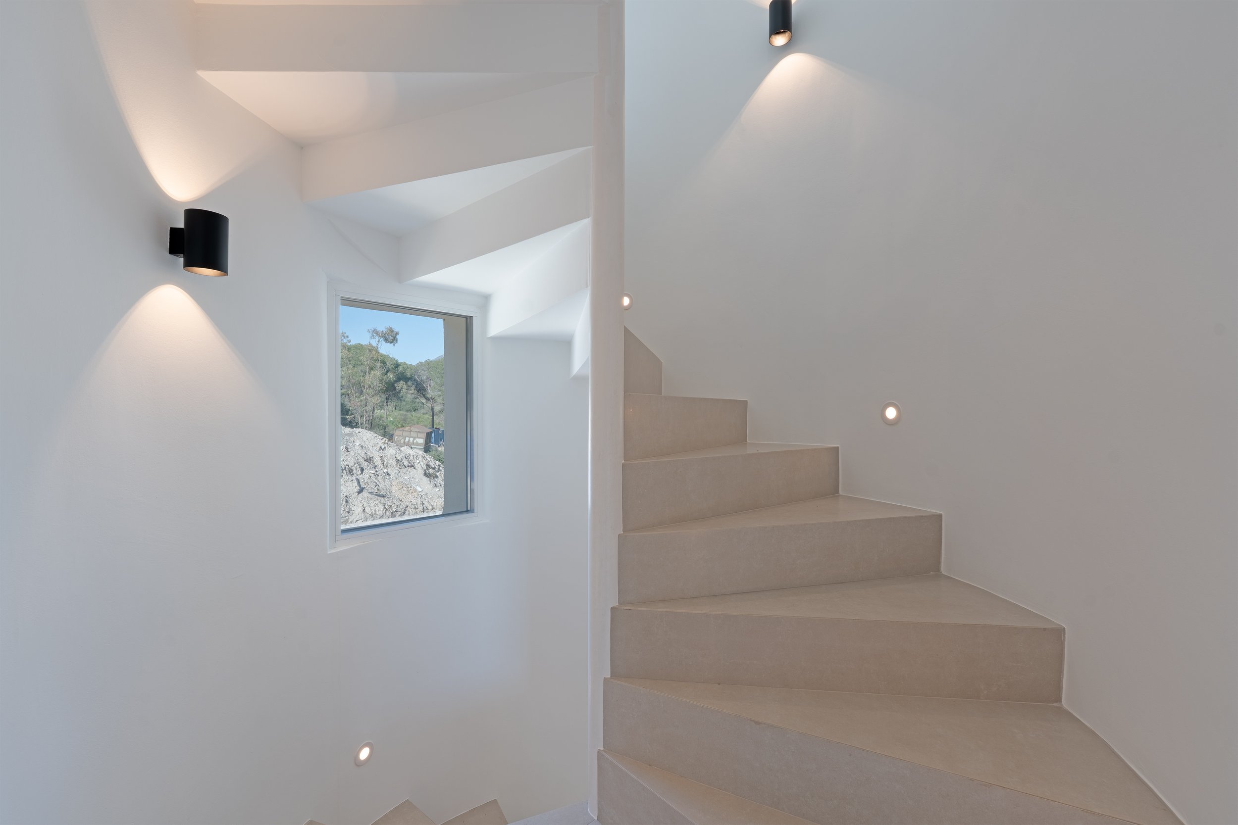 Modern interior staircase with beige steps, white walls, black wall-mounted lights, small circular lights, and a window showing an outdoor rocky landscape with trees.