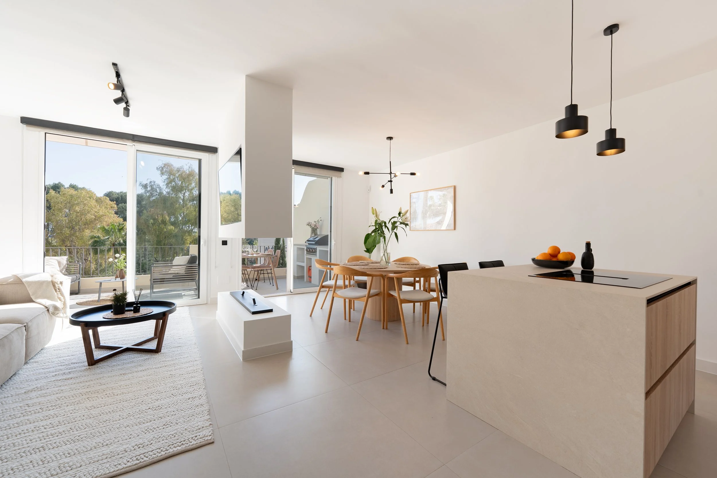 Modern open-concept living and dining area with white walls, large windows, a beige sofa, a black coffee table, a round dining table with wooden chairs, and a minimalist kitchen island with black pendant lights.