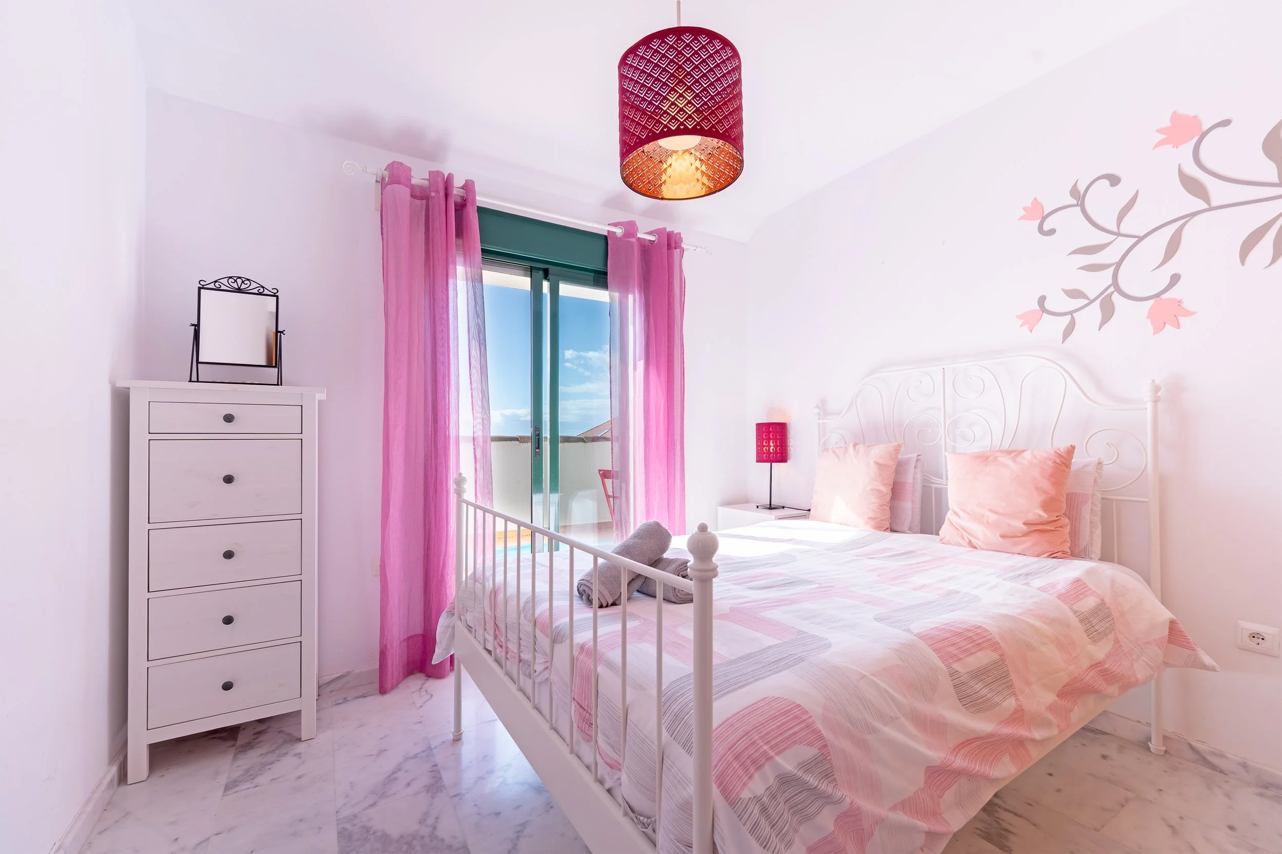 Bright bedroom with white walls, pink curtains, a white metal bed with pink pillows and a colorful bedspread, a white dresser with a small mirror, a pink table lamp, and a ceiling light with a pink lampshade. There is a sliding glass door leading to 
