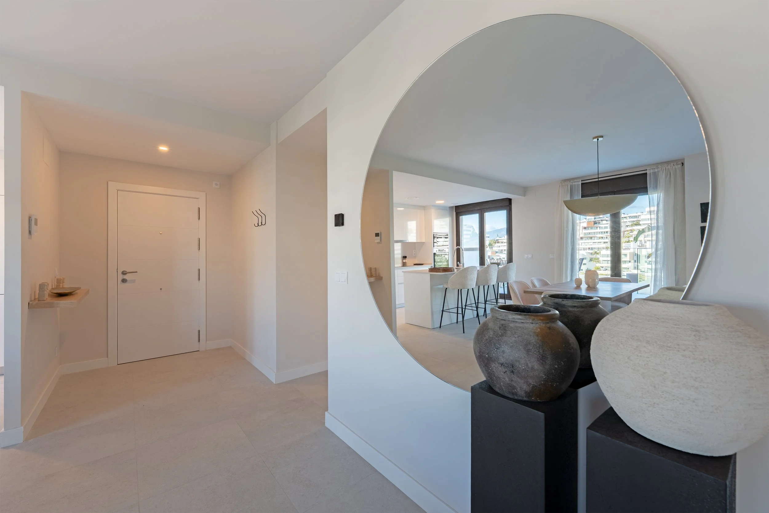 Modern apartment entryway with a view into the living and dining area, featuring decorative vases, a large round mirror, and balcony doors with a city view.