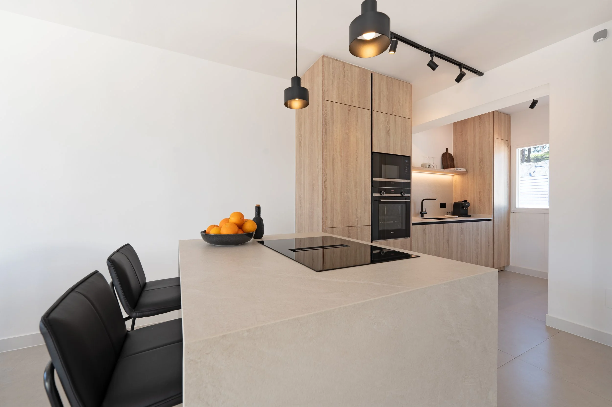 Modern kitchen with light wood cabinets, black built-in ovens, black bar-stool chairs, a white island with a fruit bowl and bottle, and track lighting on the ceiling.