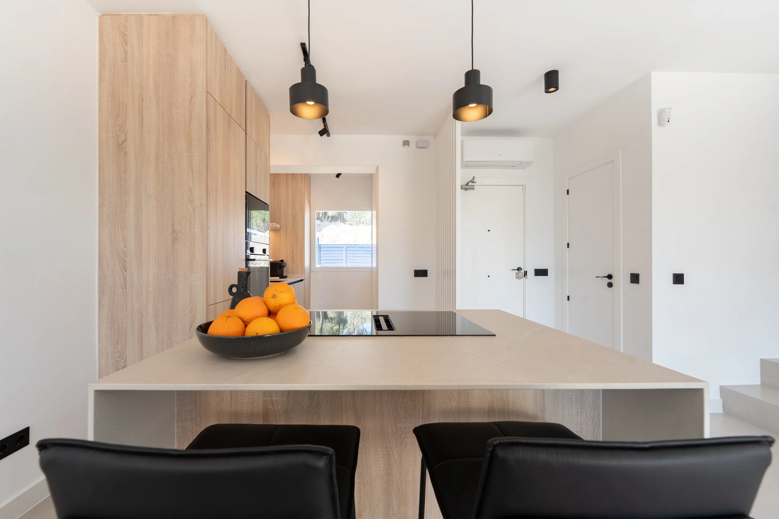 Modern kitchen with a beige island, black chairs, wooden cabinets, black pendant lights, black stove, bowl of oranges, white walls, and doors.