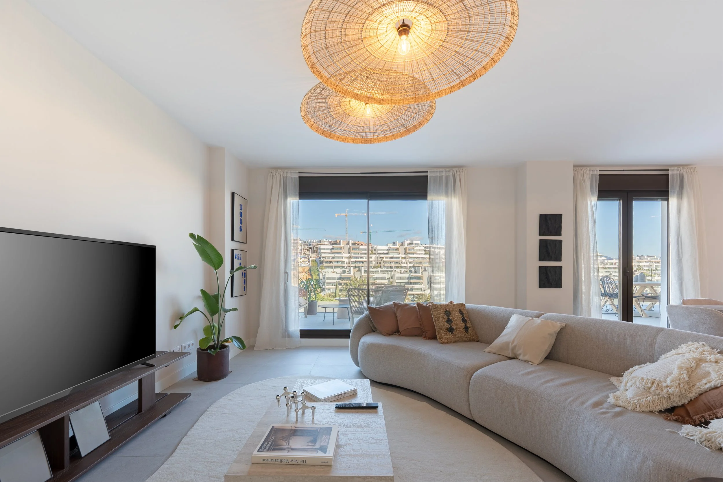 Bright living room with large windows and balcony doors, beige curved sofa with pillows, potted plant, wooden TV stand with a large flat-screen TV, ceiling lights with woven rattan shades, white walls with framed artworks, and a round rug.