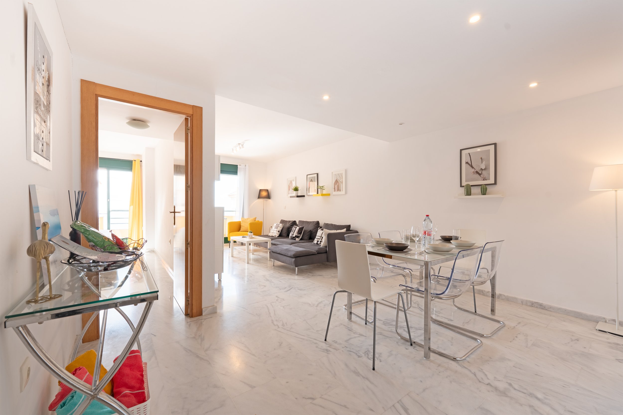 Interior of a modern, bright living and dining area with white walls and marble flooring, furnished with a dark gray sofa, a yellow armchair, a glass dining table with transparent chairs, and decorative wall art.