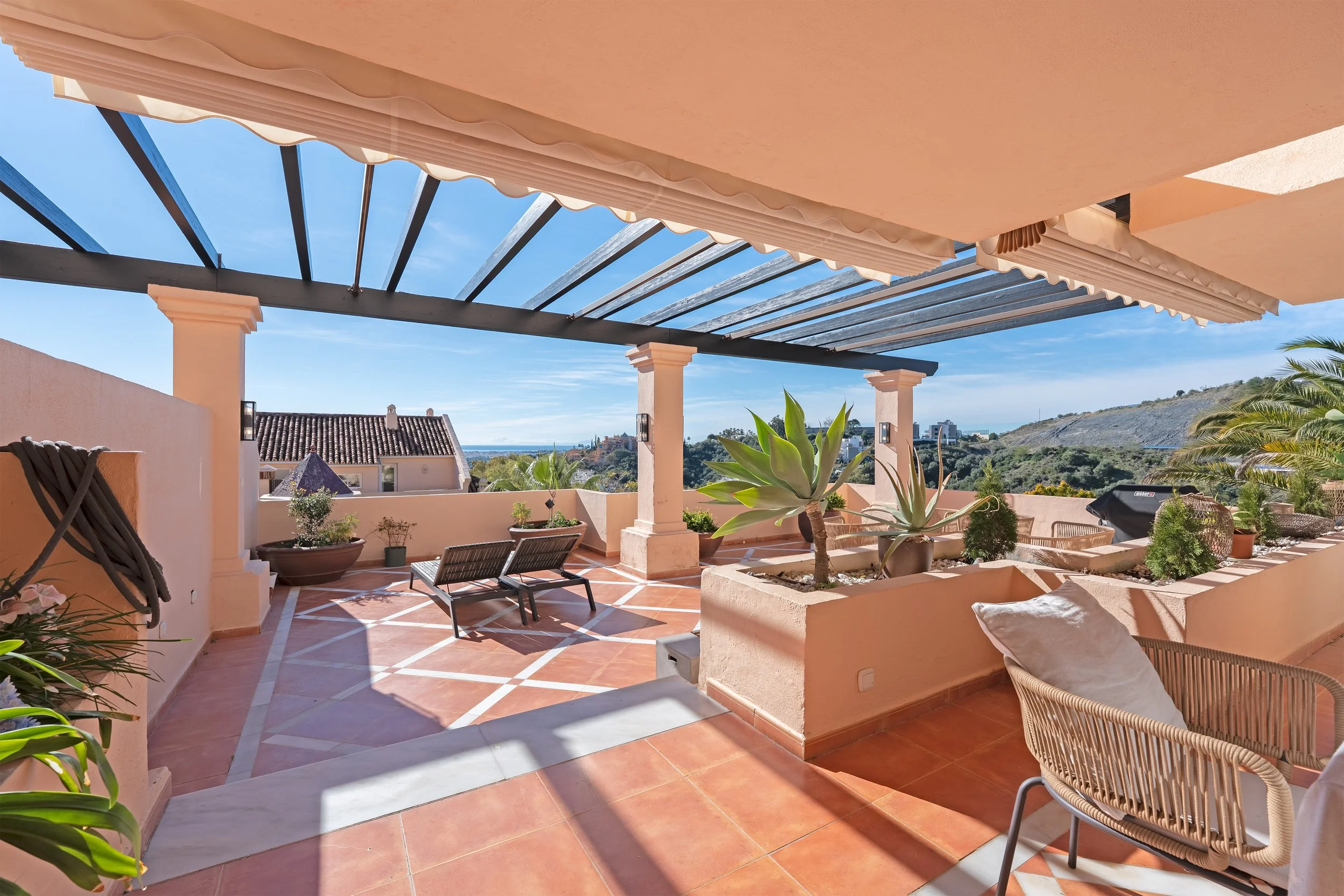 Large outdoor terrace with terracotta flooring, potted plants including two large succulents, two lounge chairs, and a wicker chair with a pillow, overlooking neighboring houses and distant hills under a partly cloudy sky.